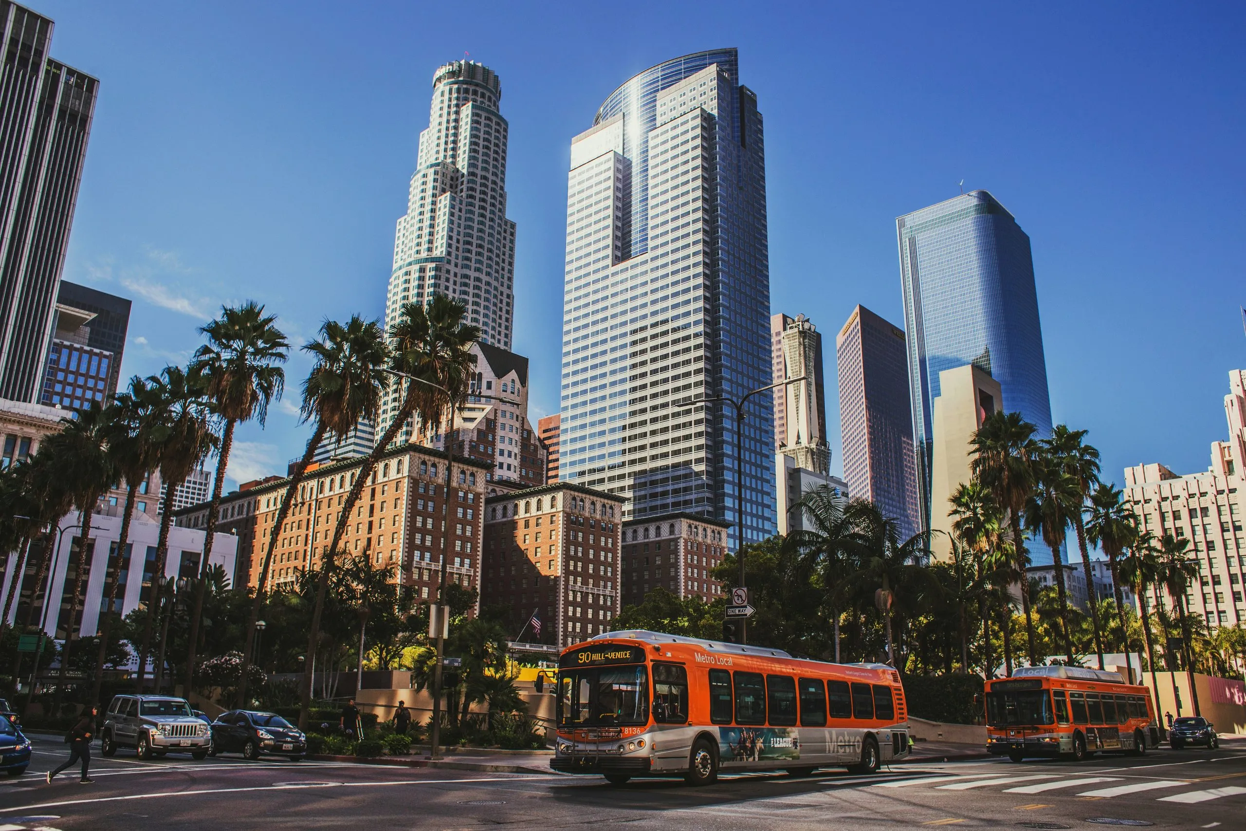 Glass downtown towers rising behind palm trees, an orange bus crossing the junction below under a clear blue sky