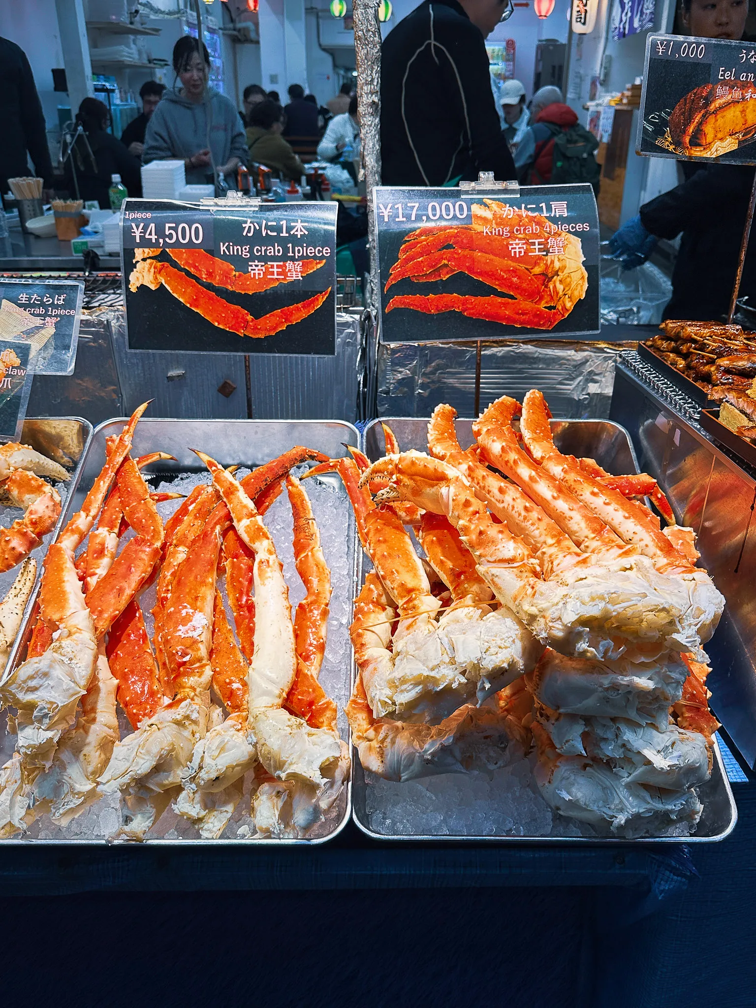 Trays of king crab legs laid on ice at a market stall, handwritten price cards propped above them