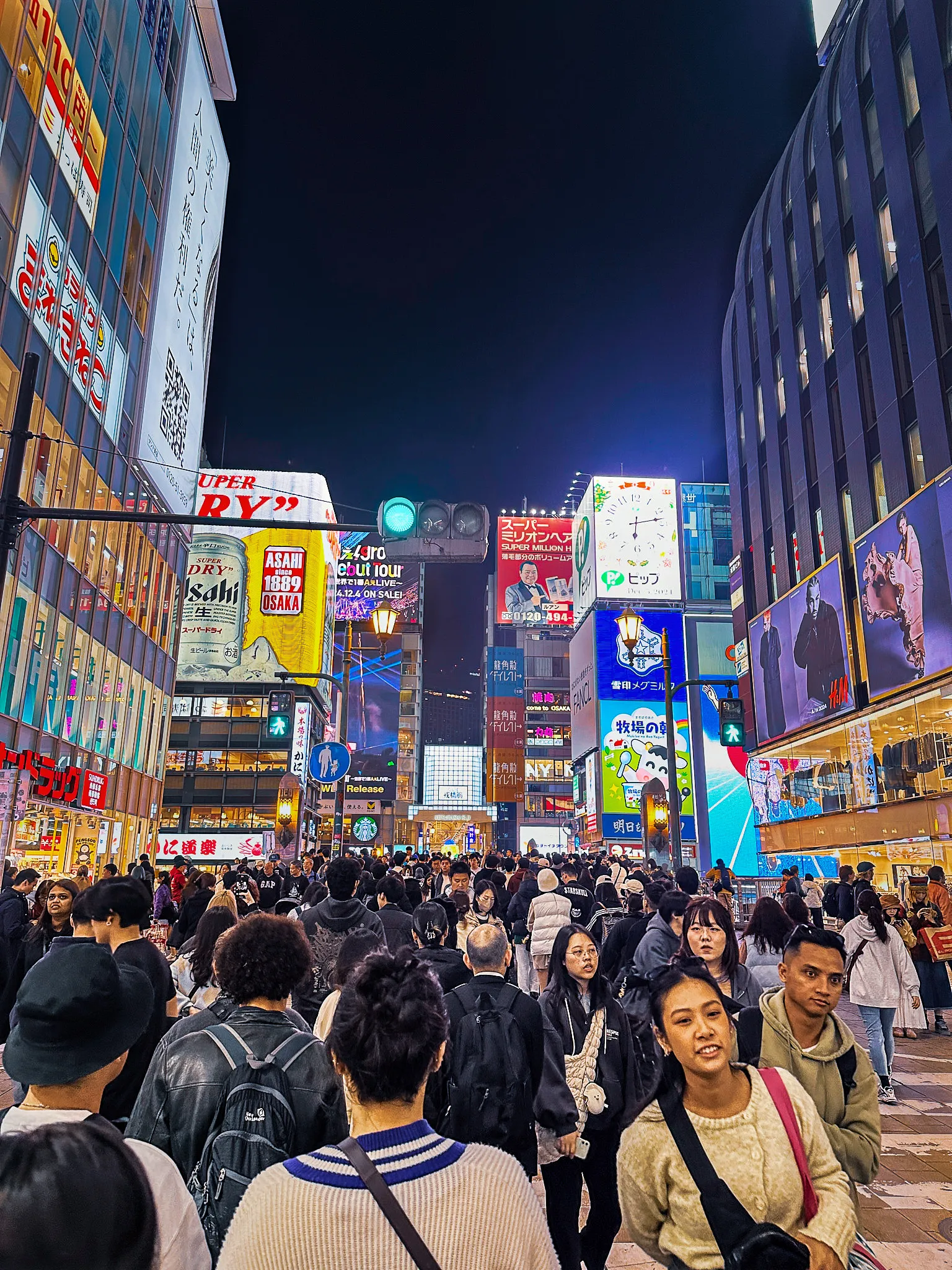 A packed street at night under stacked neon signs and video billboards, the crowd filling the road from side to side