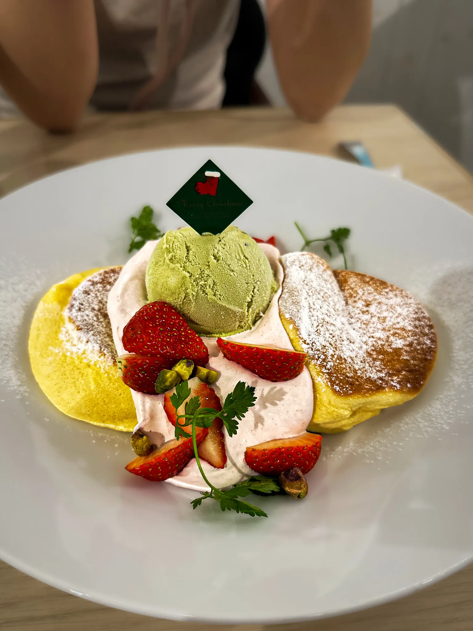 Two souffle pancakes dusted with sugar on a white plate, with a scoop of matcha ice cream, strawberries, cream and pistachios