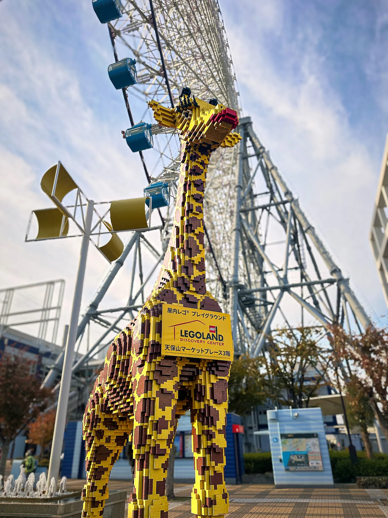 A giraffe built from LEGO bricks standing outside an entrance, a ferris wheel rising behind it against a pale sky