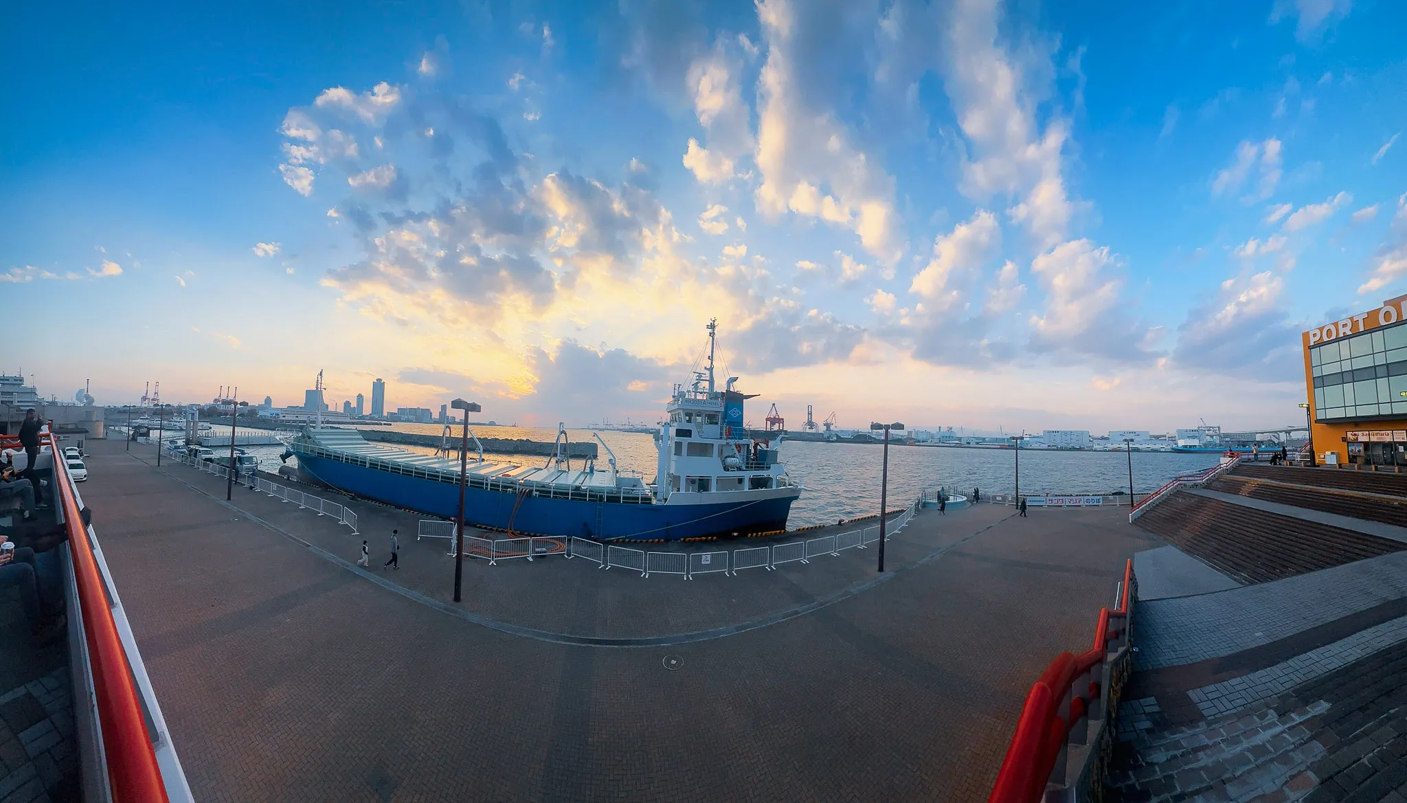 A wide panorama of the harbour at sunset, a blue cargo ship moored along the promenade and cloud lit gold above the cranes beyond
