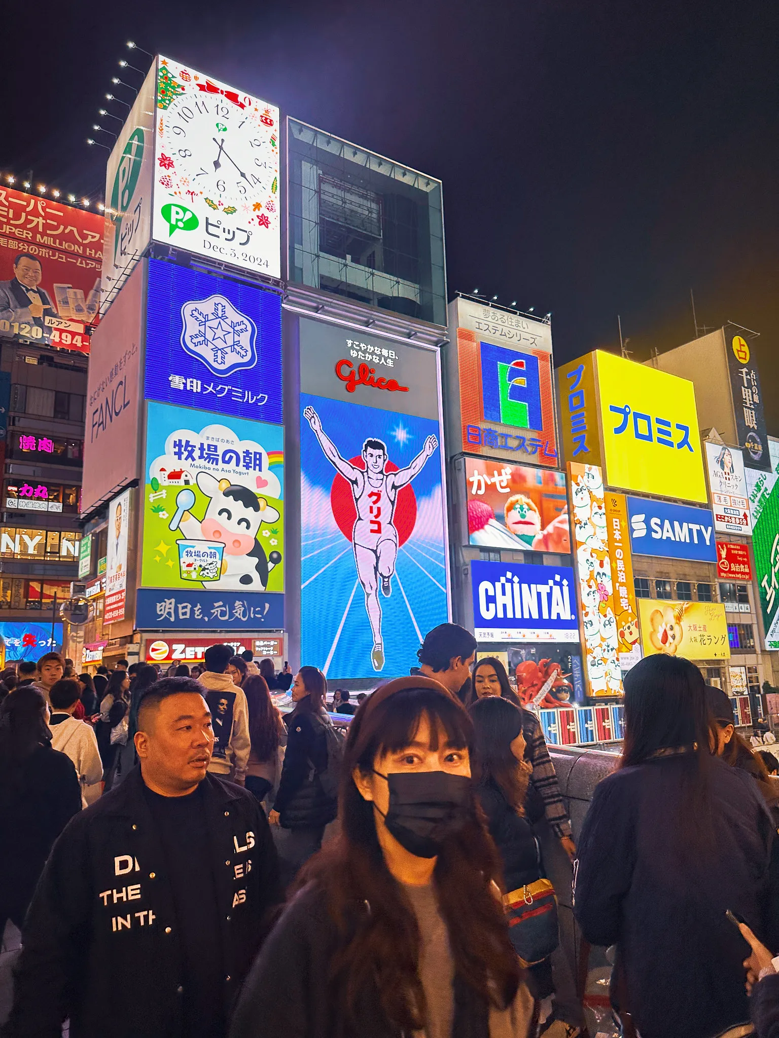 Dotonbori at night under stacked billboards, the Glico running man lit up in the middle and a clock face beside it, the crowd pressing past below