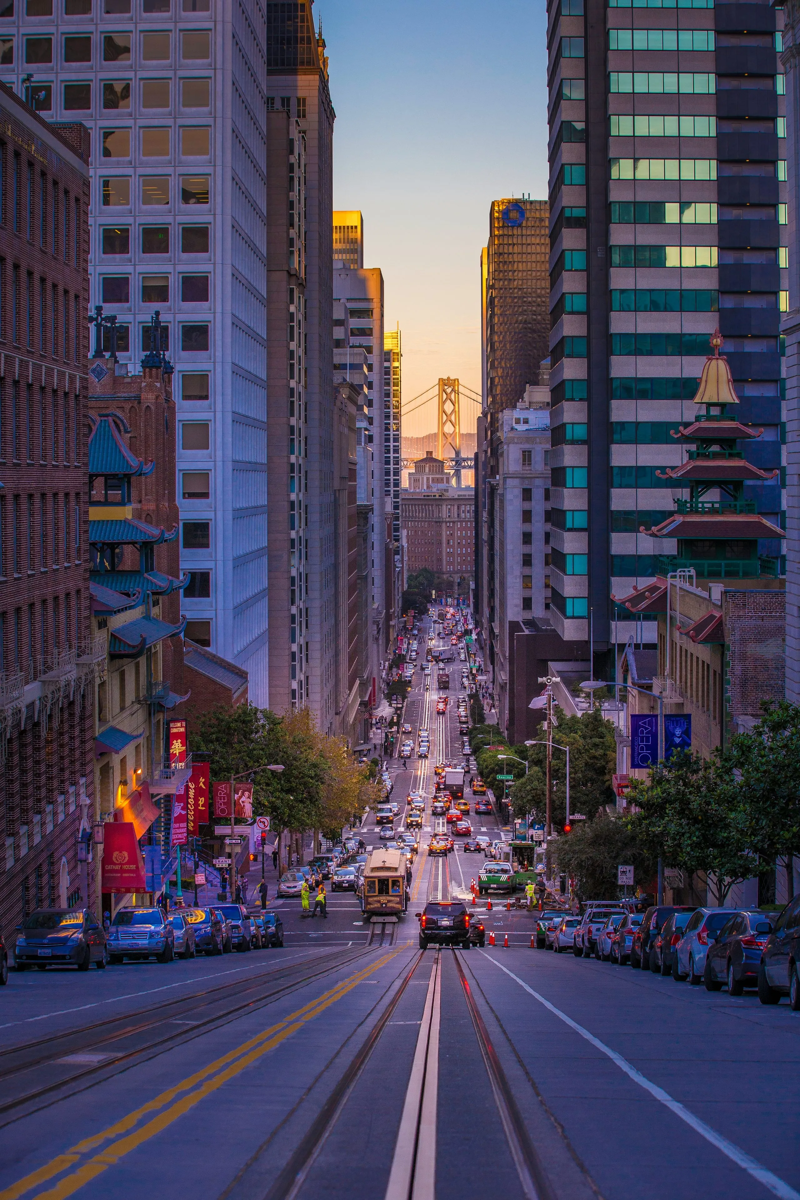 California Street's tracks running downhill between towers toward the Bay Bridge at dusk, shopfronts glowing on both sides
