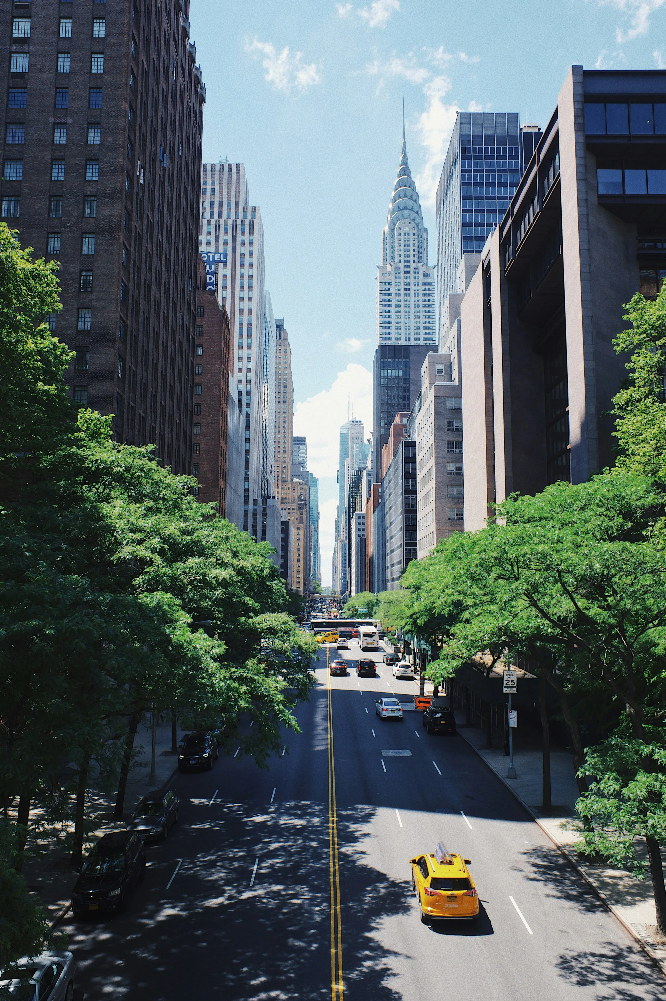 Looking down a tree-lined avenue toward the Chrysler Building's spire, a single yellow cab in the road below