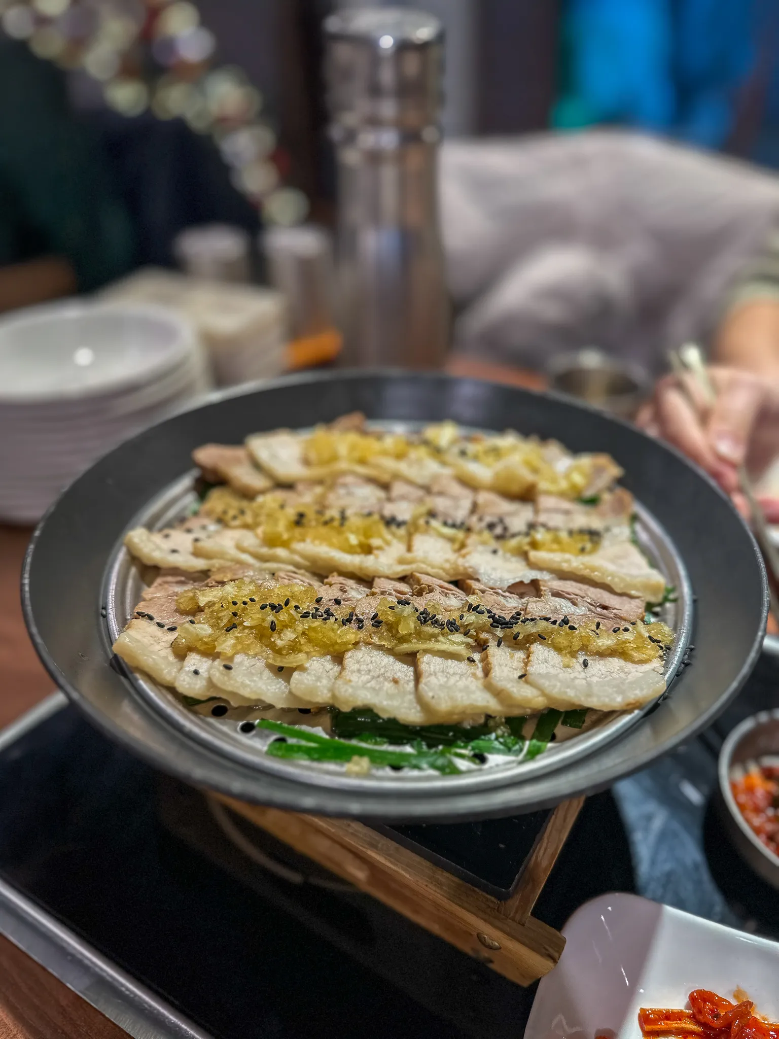 Slices of pork belly laid in rows on a domed grill pan over spring onions, a garlic and ginger relish and black sesame over the top, kimchi and dishes around the edge