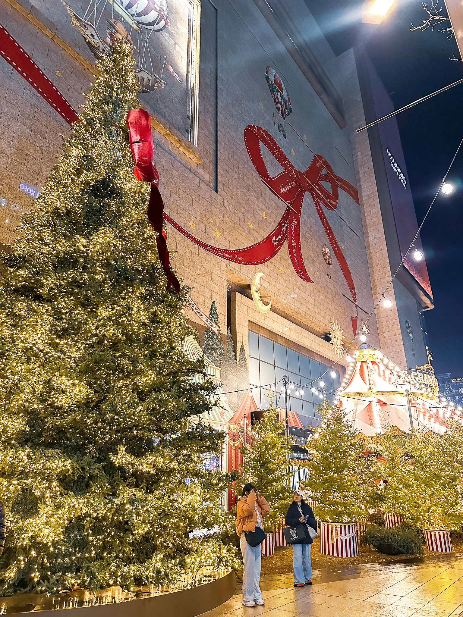 A tall lit Christmas tree in front of a department store wrapped in a painted red ribbon and bow, a carousel and striped tents glowing behind