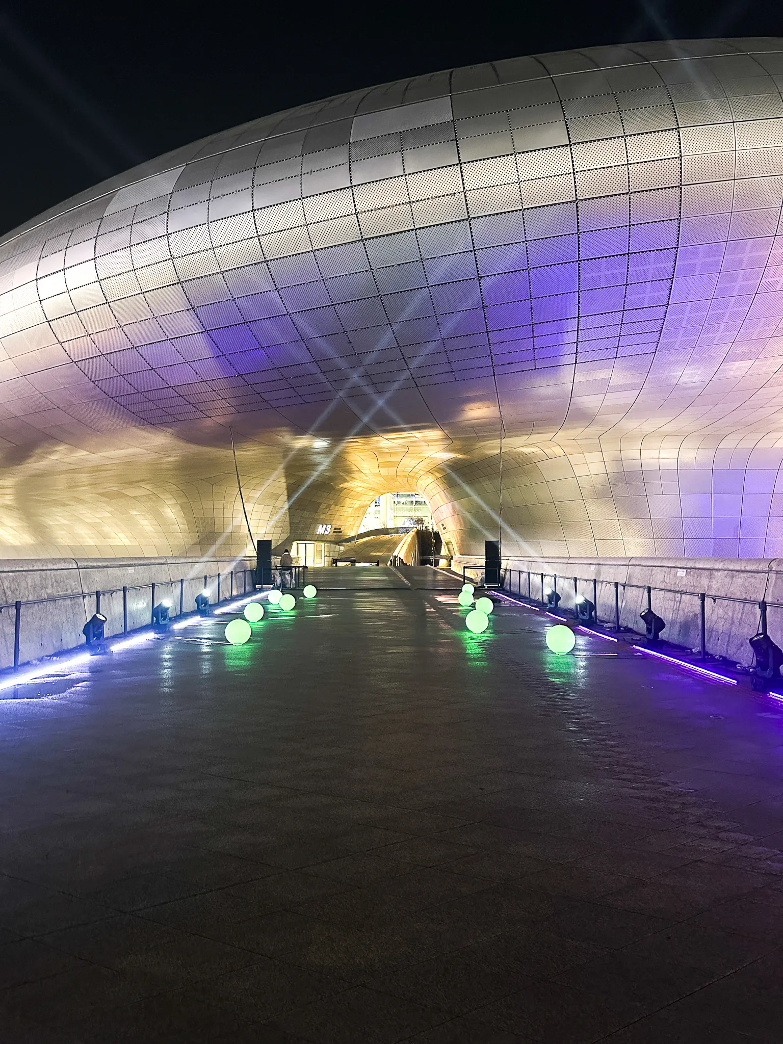 The curved silver skin of a building arching overhead at night, lit purple and gold, a wet ramp running underneath it lined with glowing spheres