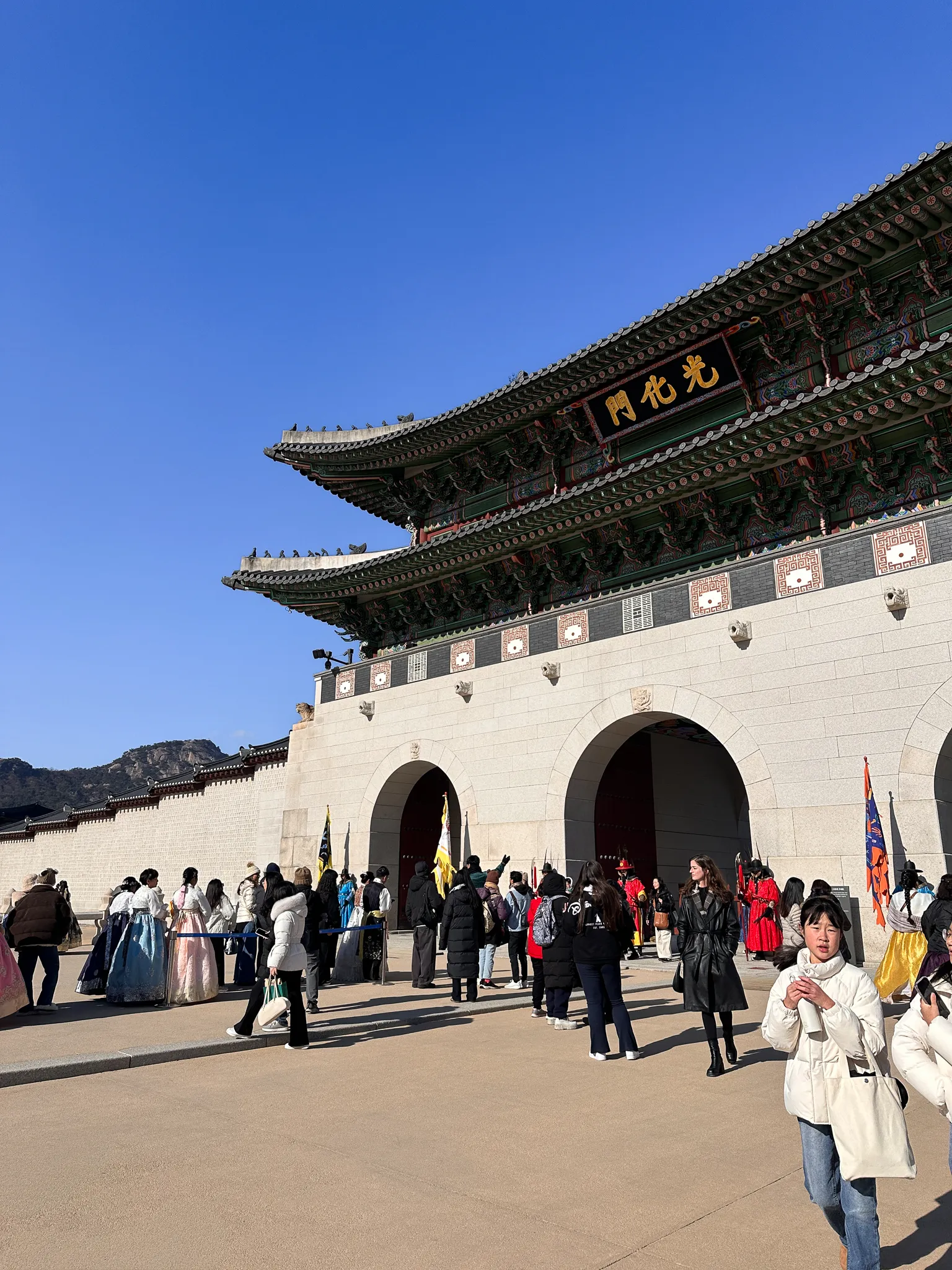 Gwanghwamun from an angle with its gold sign lit by low sun, a line of visitors in rented hanbok along the wall and a guard in red standing by the arch