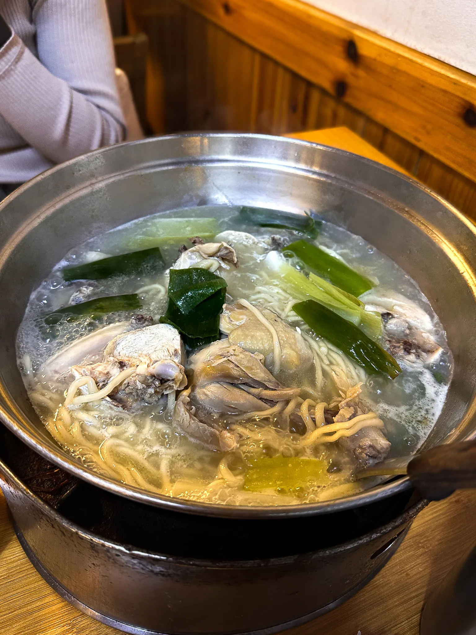 A wide metal pot of chicken pieces simmering in clear broth with leek, noodles and rice cake, set on a burner at the table