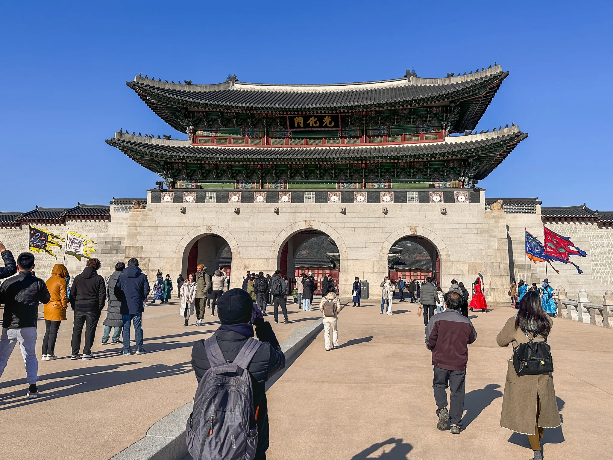 The two-storey Gwanghwamun gate straight on, its three arched openings open and crowds crossing the plaza in front, banners standing either side