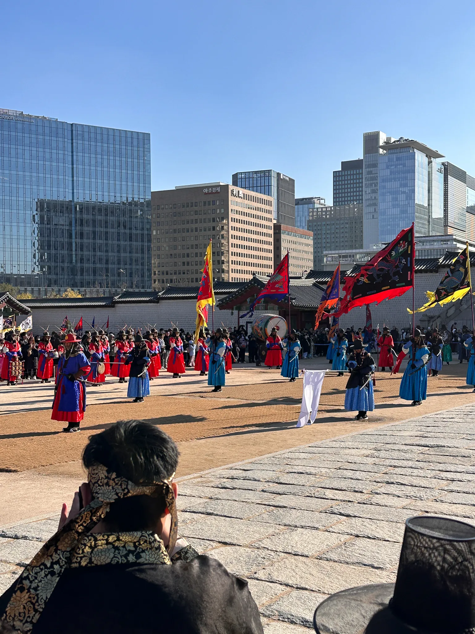 Ranks of guards in blue and red crossing the plaza with tall painted banners, a drum among them, the palace wall in front and office towers rising behind