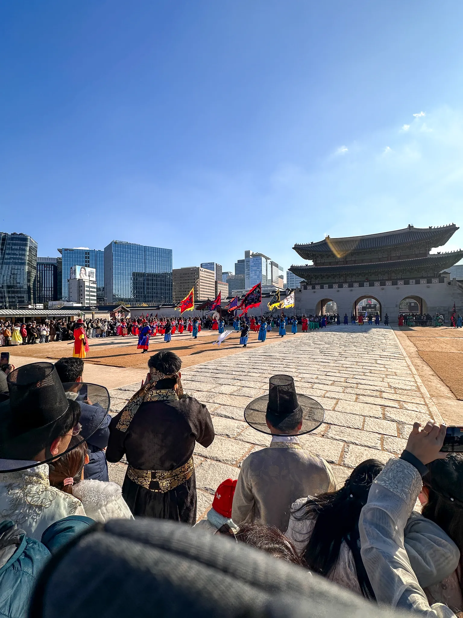 The guard ceremony in the plaza seen over the shoulders of people in hanbok and black horsehair hats, ranks in blue and red with flags, the gate and the city behind