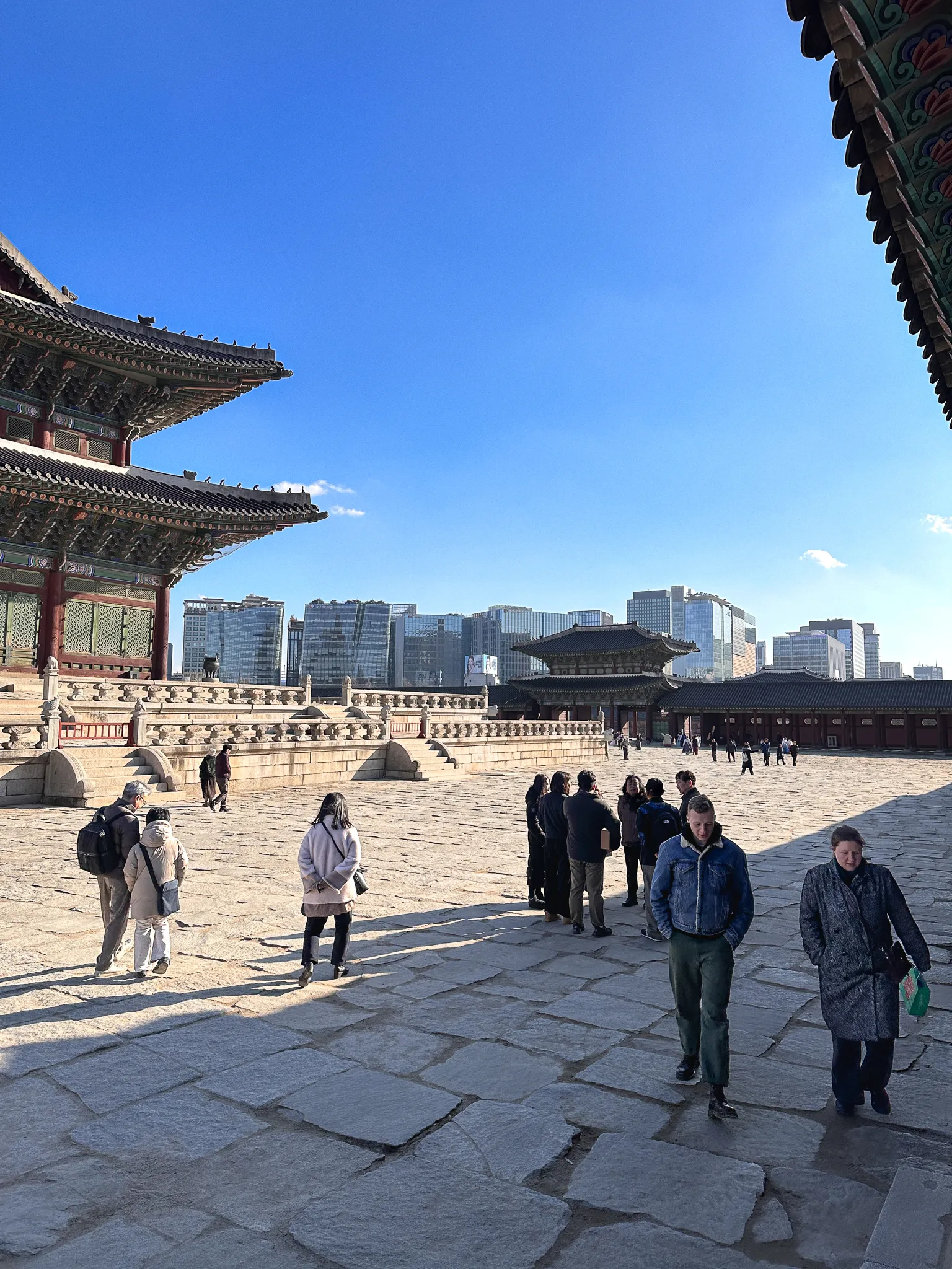 A palace courtyard of worn flagstones with painted eaves on both sides, and beyond the far wall a row of glass office towers
