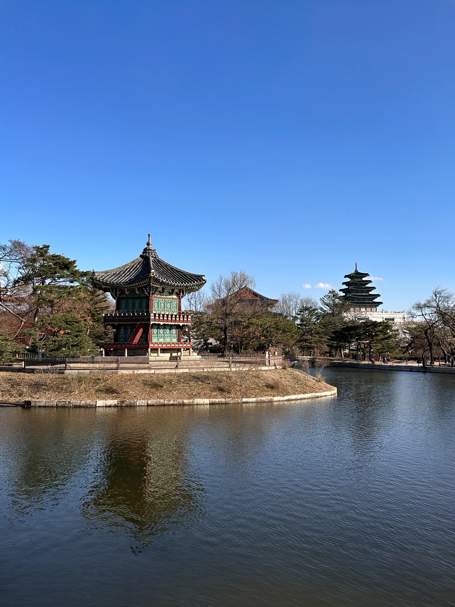 The same pavilion from further along the bank, a five-storey tower rising among the trees behind it and the pond flat and brown in front