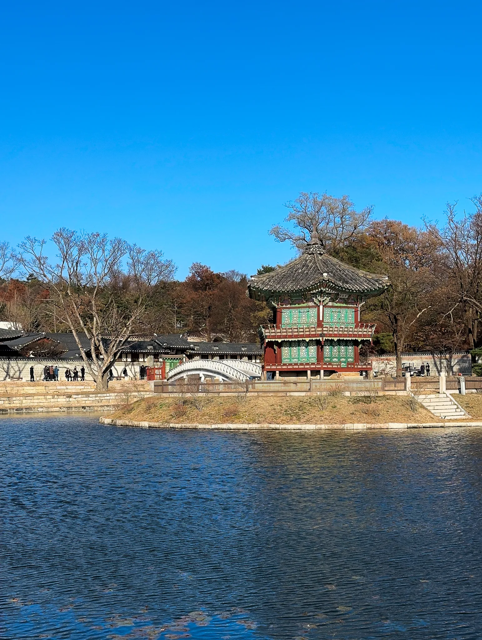 A small two-storey hexagonal pavilion on its own island, a white arched bridge reaching it from the left, bare winter trees behind and hard blue sky above