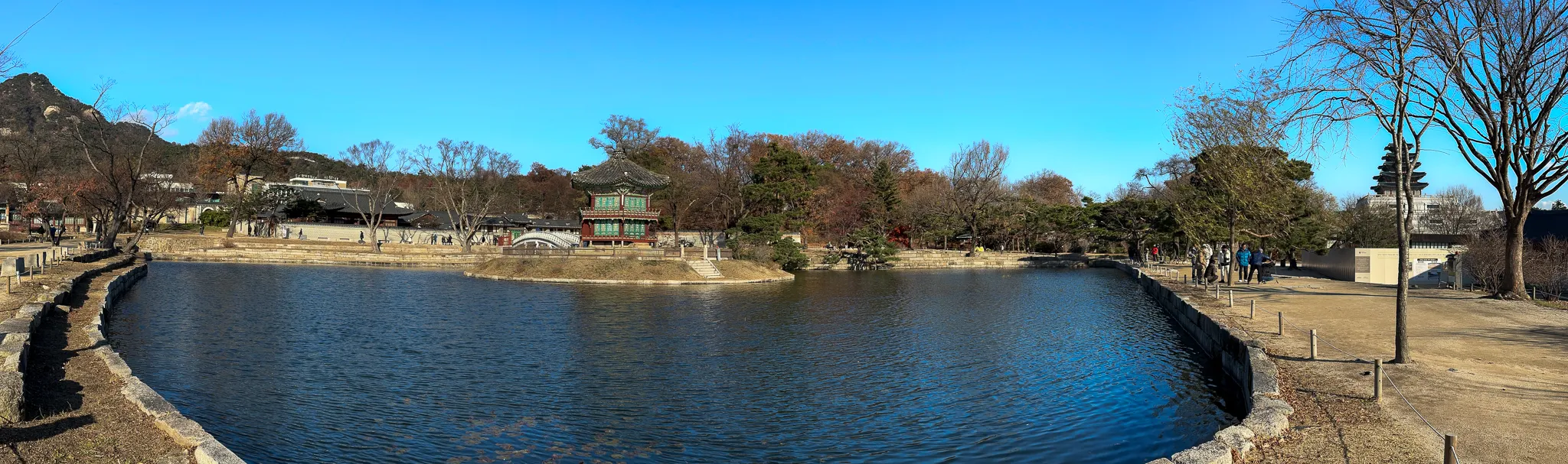A wide panorama of the palace pond, the pavilion small on the far bank between bare trees, a granite kerb running around the near edge and hills beyond