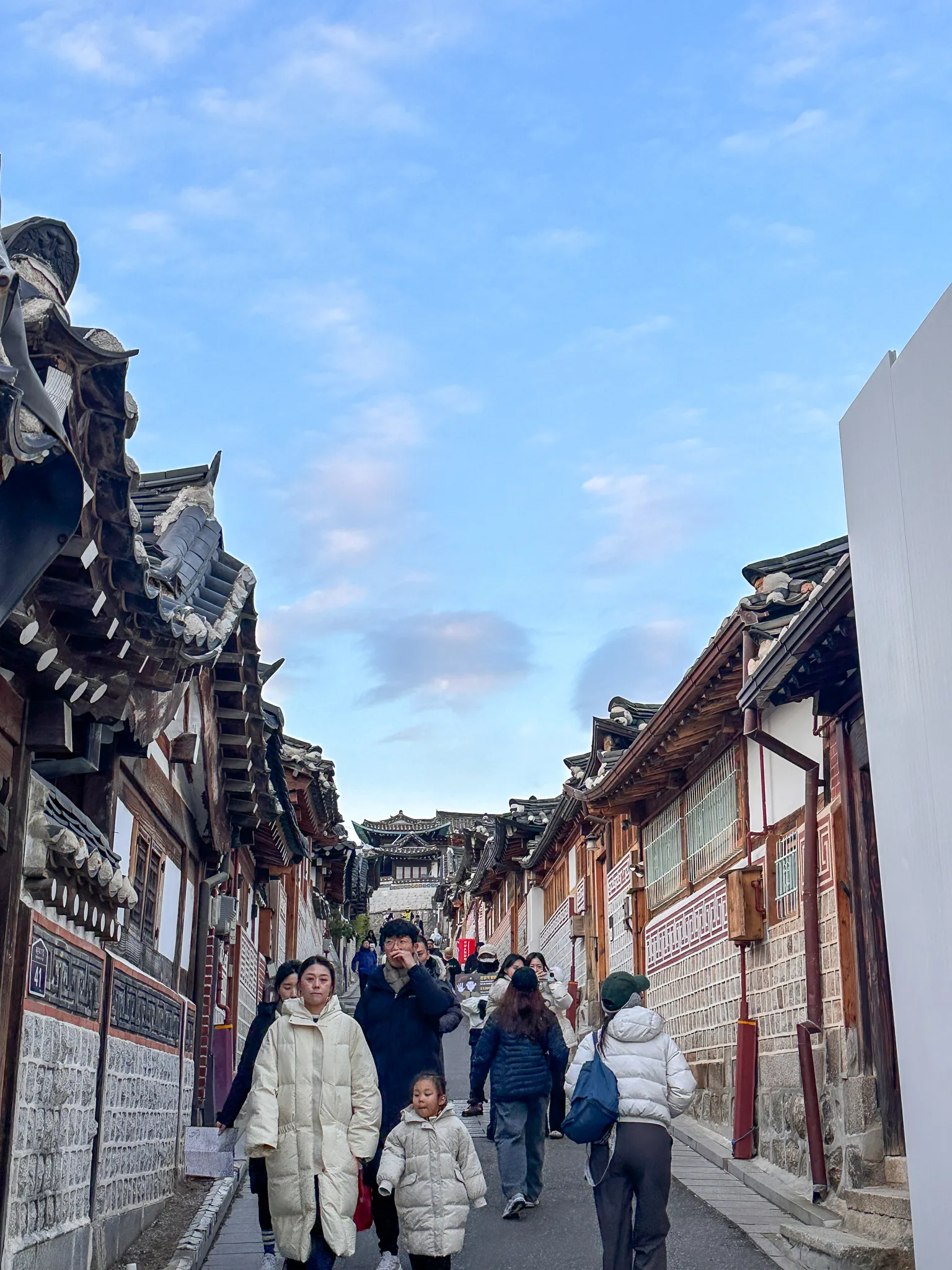 A narrow lane between hanok walls and tiled eaves, people walking down it in winter coats and a gate standing at the top under a pale blue sky