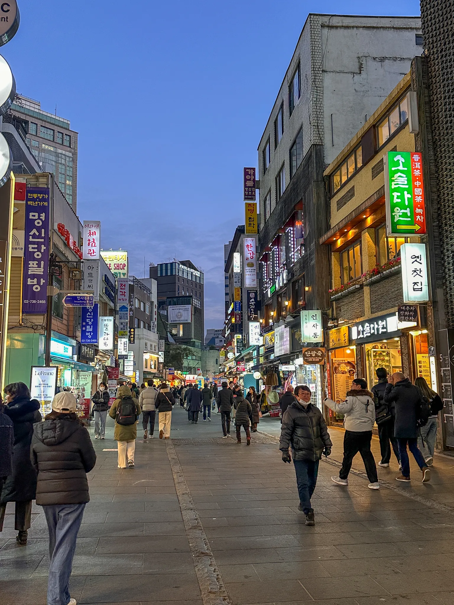 A pedestrian street at dusk under a deep blue sky, hand-painted signboards stacked up both sides, teahouses and shops lit, everyone in heavy coats