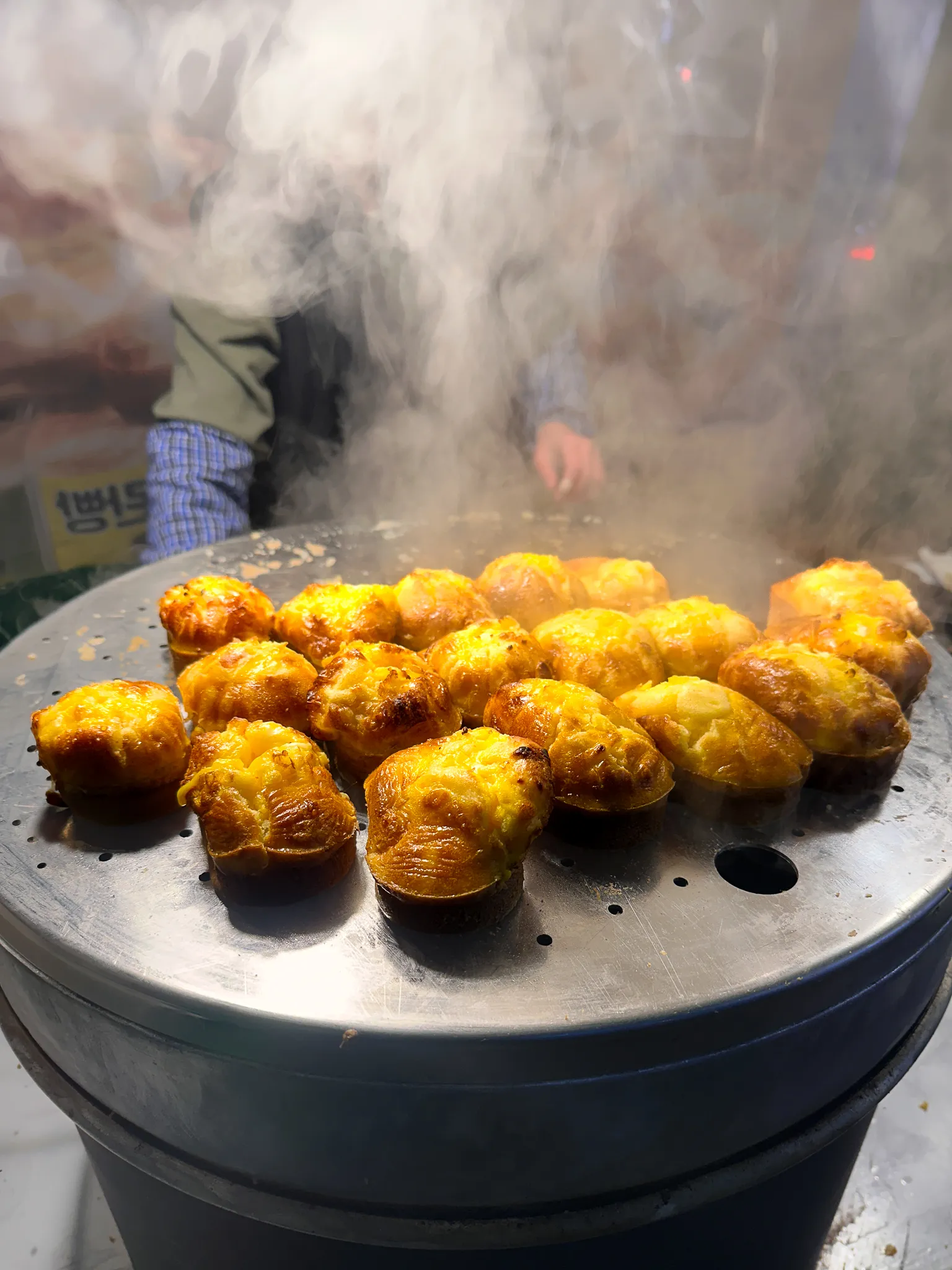 A row of egg breads browning on a street vendor's steel steamer, steam pouring off them and the vendor's hands just visible behind