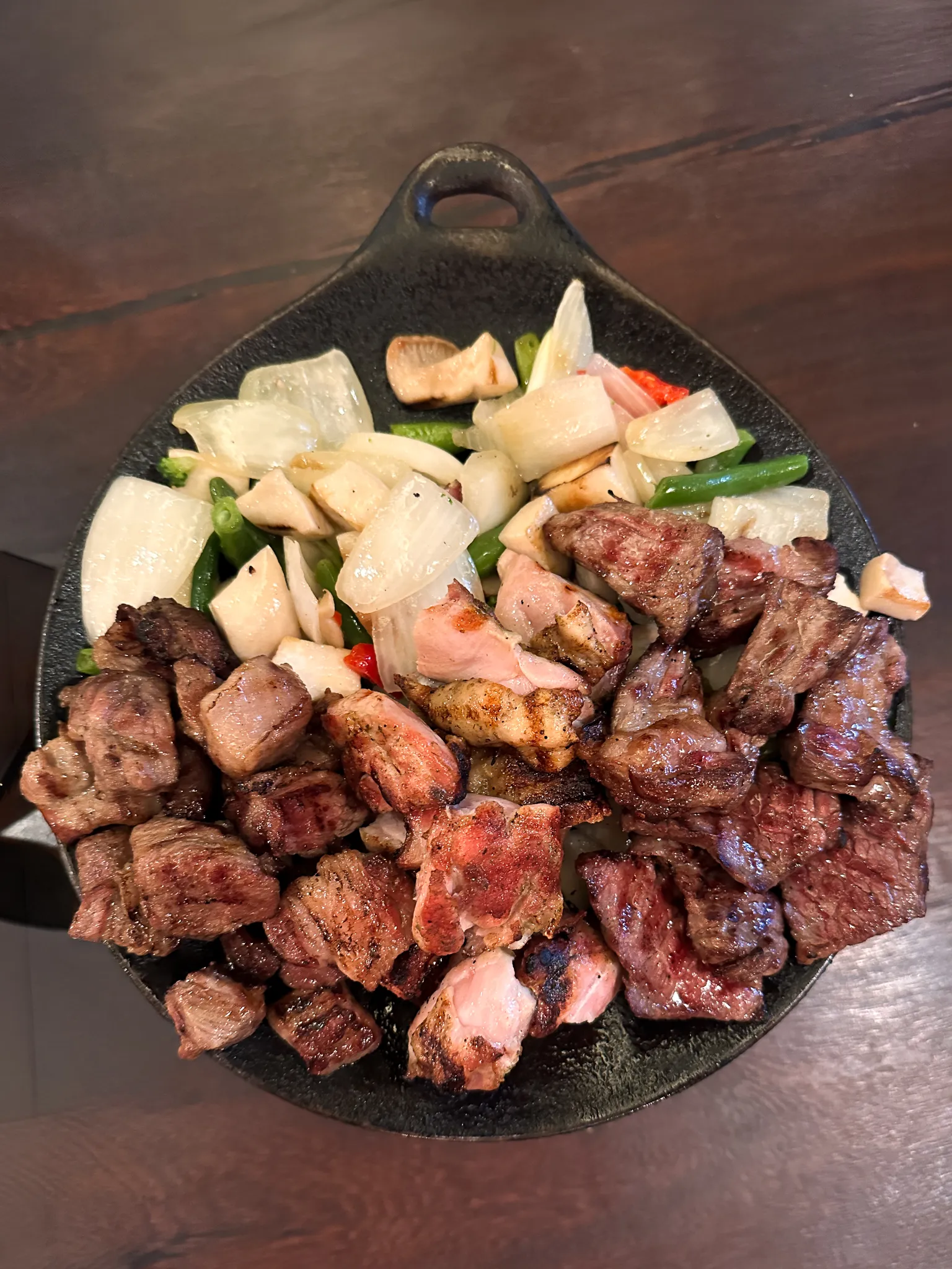 A cast-iron pan of cubed grilled beef with onion, garlic, green chilli and beans pushed to one side, on a dark wooden table