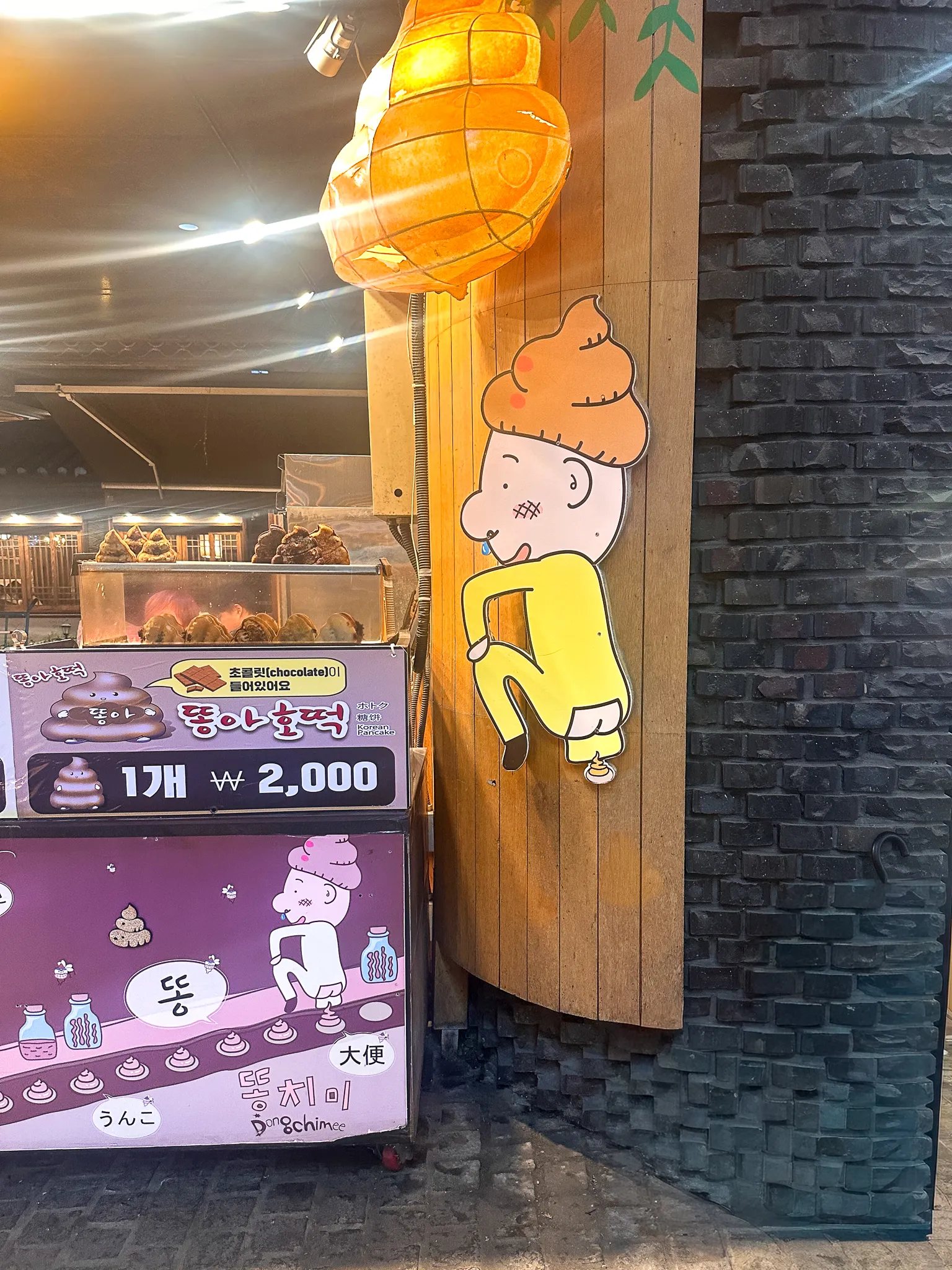 A street stall selling bread shaped like a swirl of poop, a cartoon character painted on the wooden wall beside it and a paper lantern glowing overhead, the price board reading two thousand won