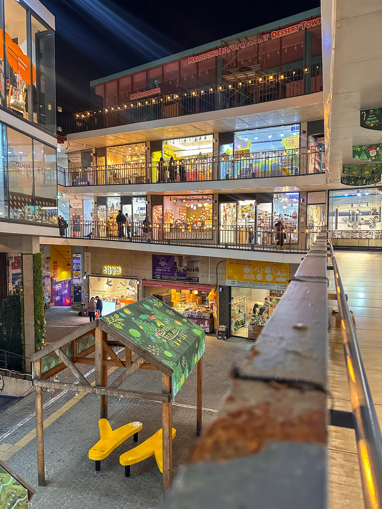 A market building at night seen from an upper walkway, shops lit on three levels around an open courtyard, a painted wooden shelter and yellow play equipment on the ground floor