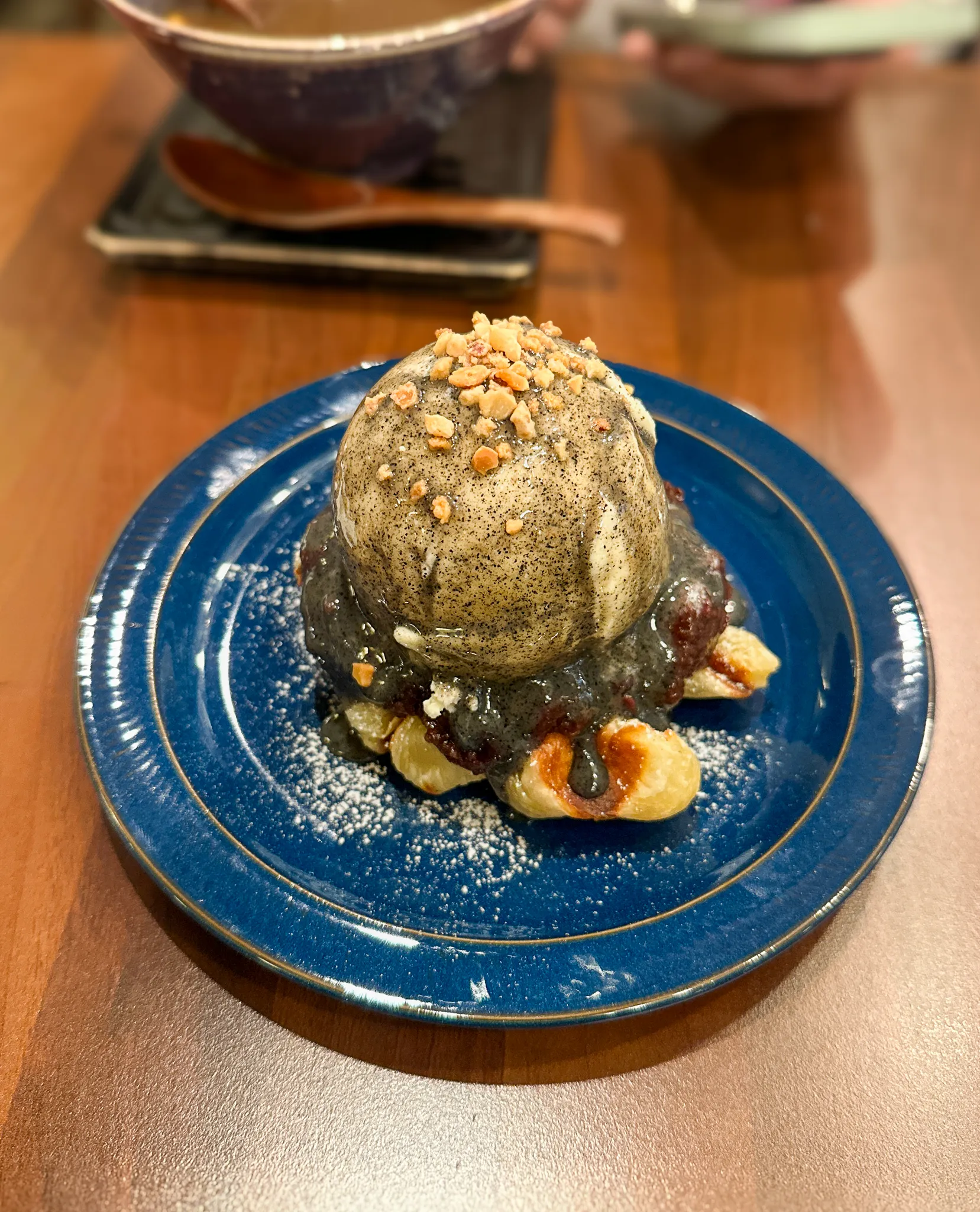 A scoop of grey-flecked ice cream over red bean paste and pieces of fried dough on a deep blue plate, crushed peanuts on top and icing sugar dusted around it