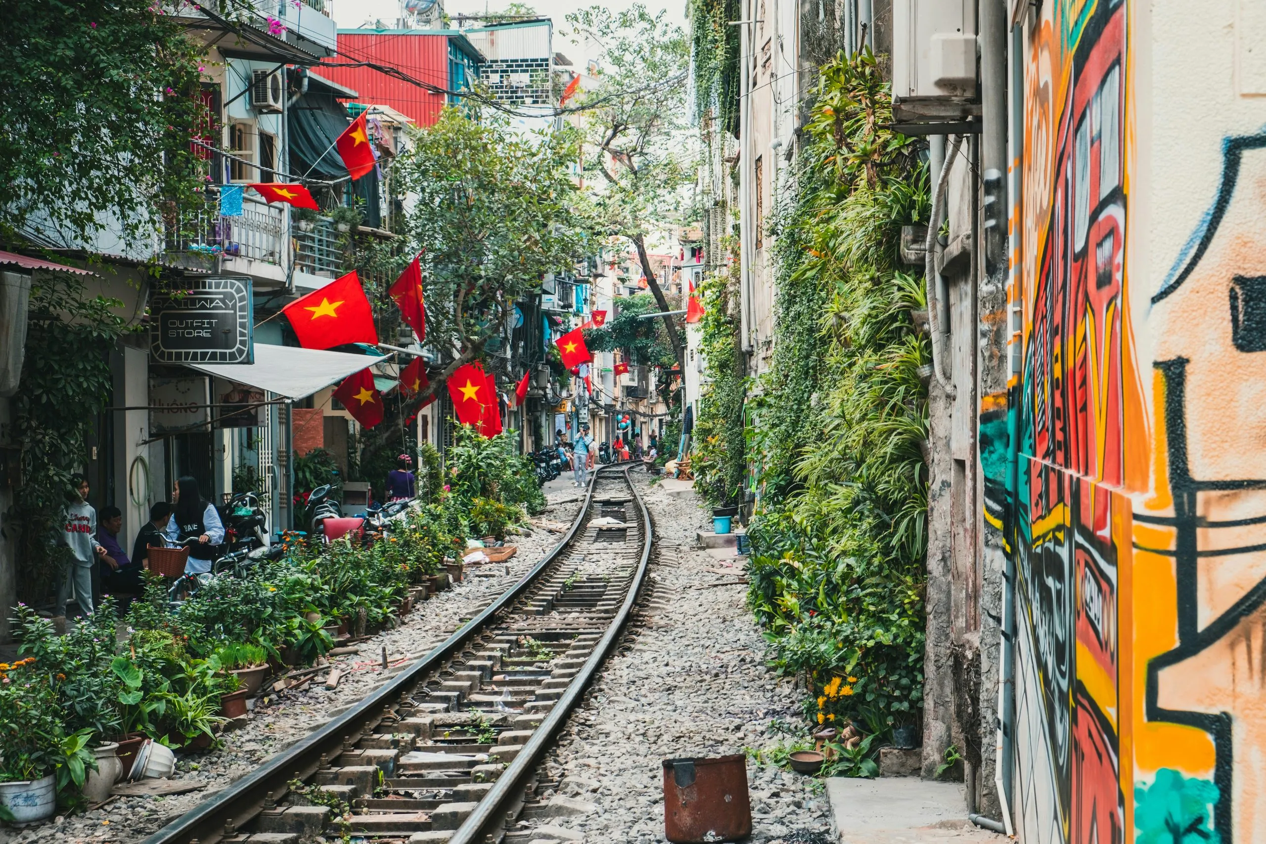 Railway tracks running between the back walls of houses on Train Street, Vietnamese flags strung overhead and a mural along the right-hand wall
