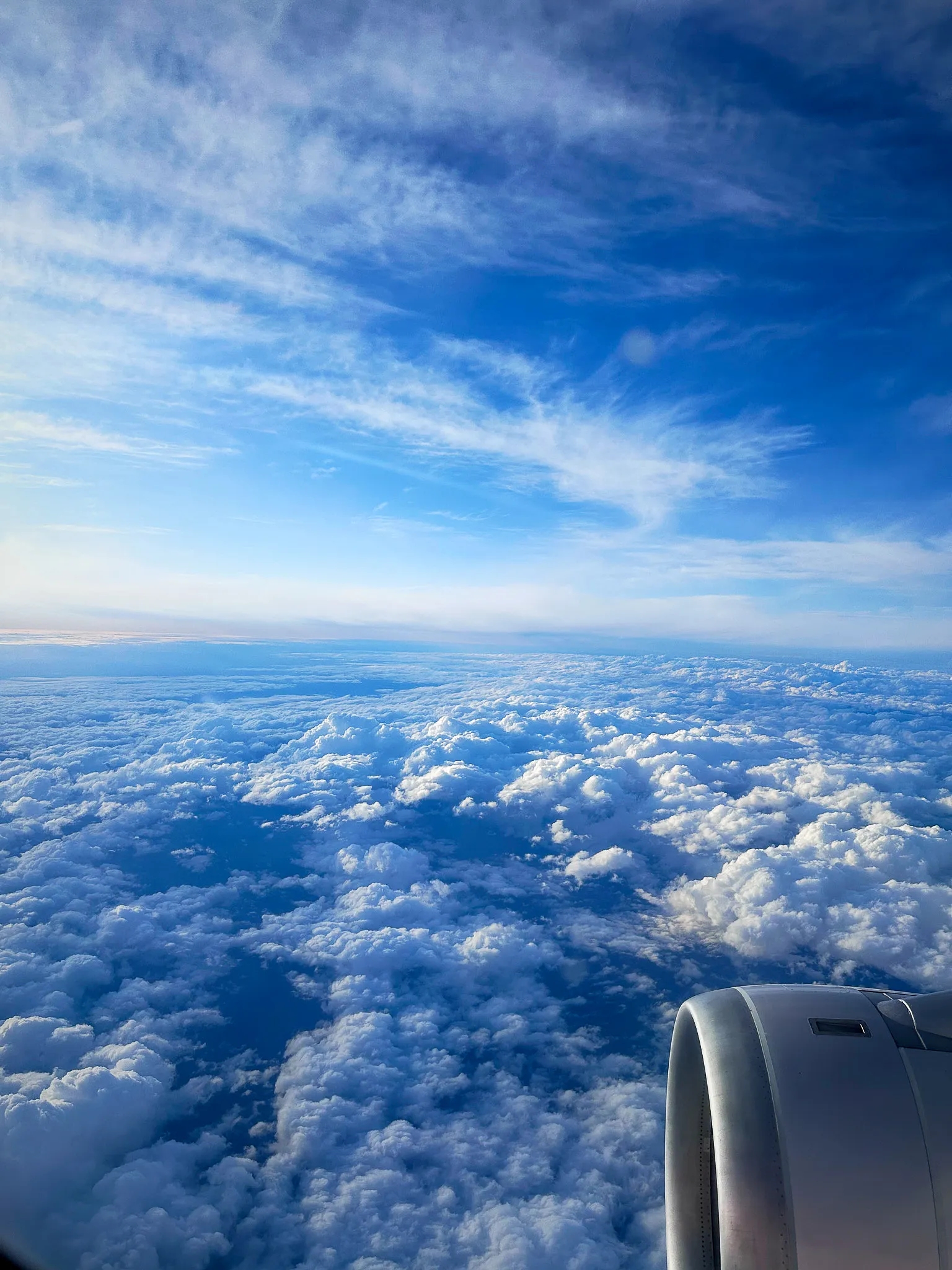 The view from a plane window: an engine at the lower right, a deck of cloud below, and wisps of cirrus across a blue sky