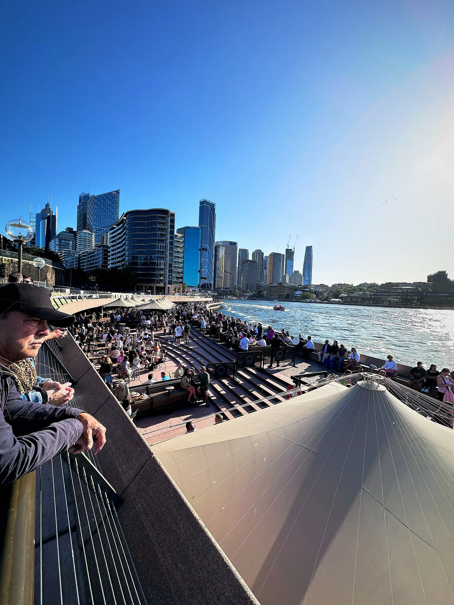 Circular Quay from above the Opera Bar's sail canopies, crowds along the waterfront and the city towers across the water, a man in a dark hat leaning on the railing in the foreground
