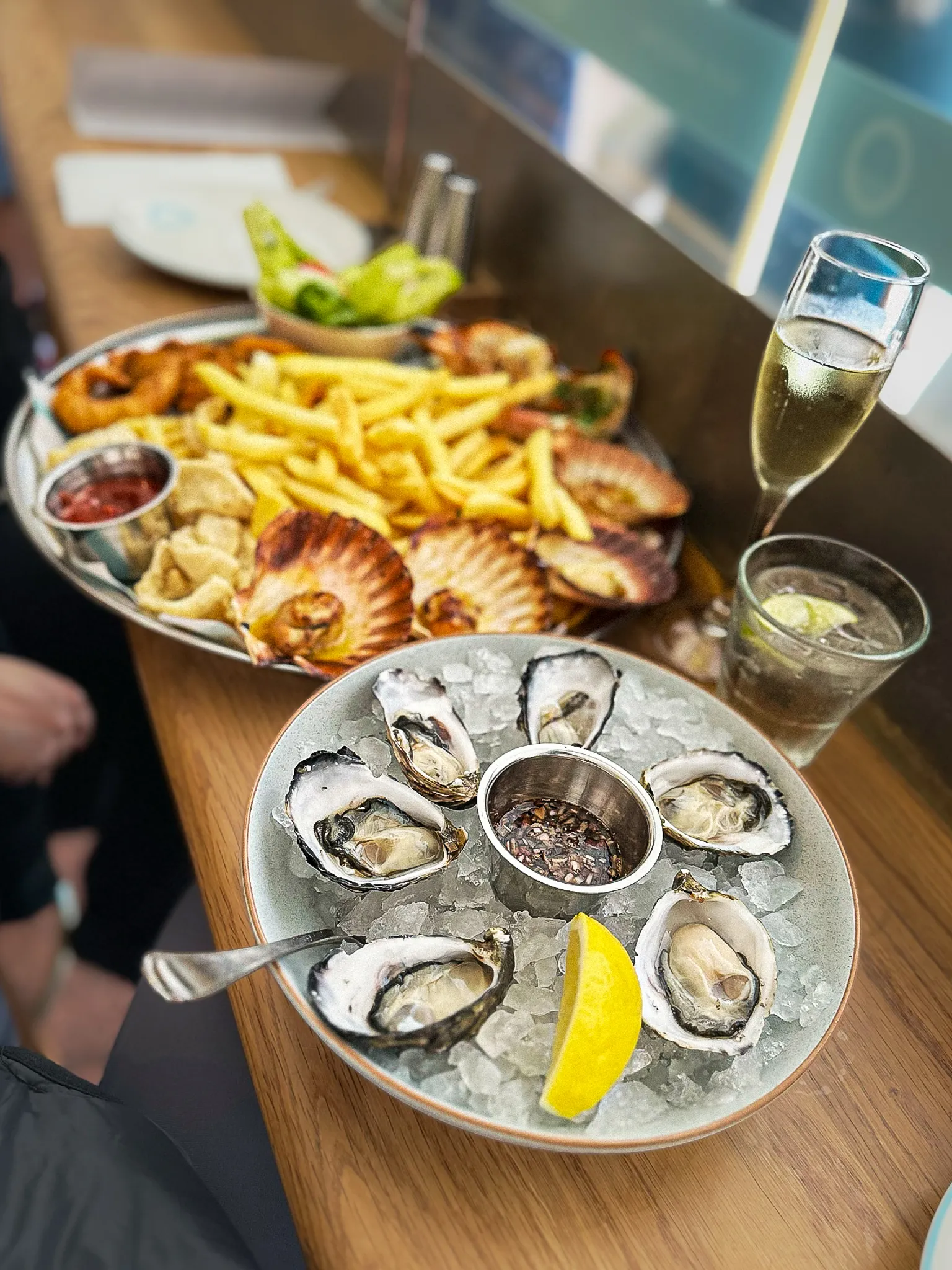 Six oysters on crushed ice around a pot of mignonette with a lemon wedge, a platter of chips, calamari and scallops behind, and a glass of sparkling wine