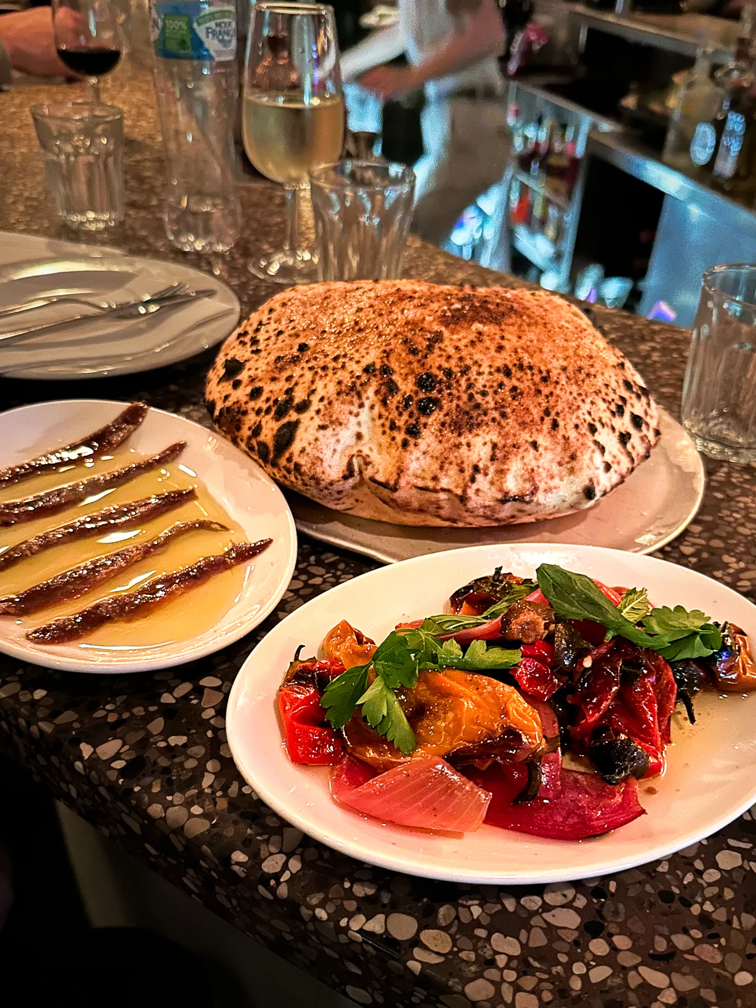 A terrazzo bar counter with anchovy fillets in olive oil, a puffed woodfired flatbread, and a plate of roast peppers and tomatoes scattered with herbs
