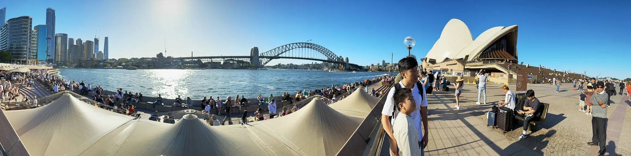 A wide panorama of Circular Quay: the city towers on the left, the Harbour Bridge across the middle, the Opera House on the right, and people gathered along the water's edge
