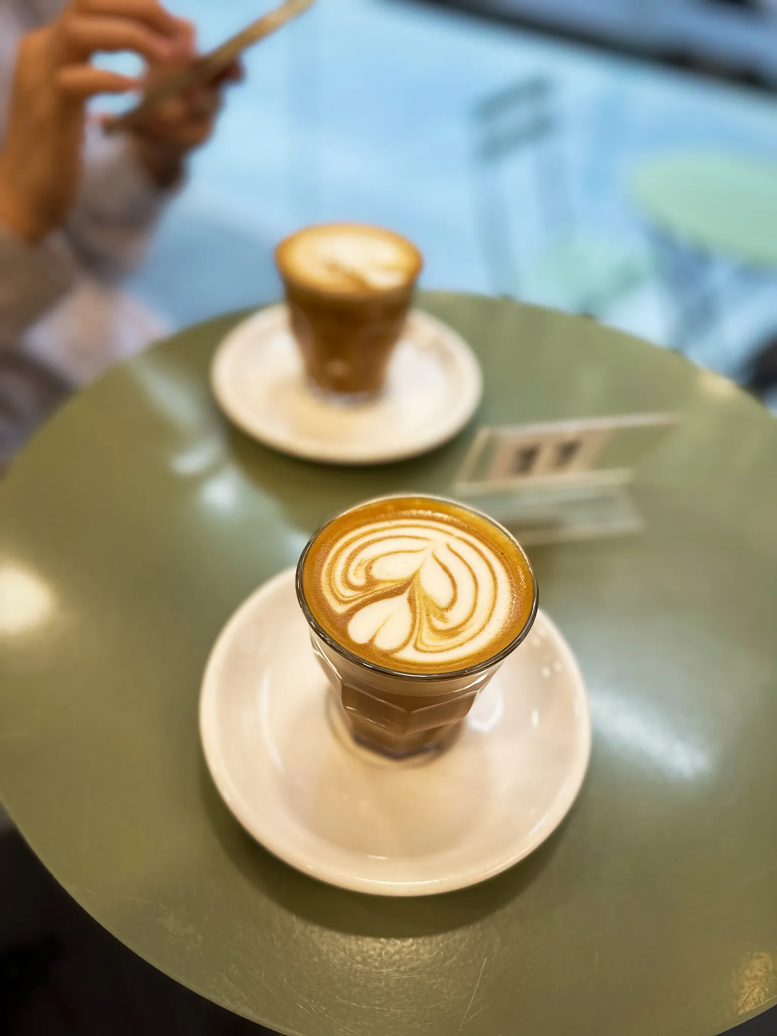 Two coffees in glasses on saucers on a sage-green cafe table, a fern poured into the foam of the nearer one