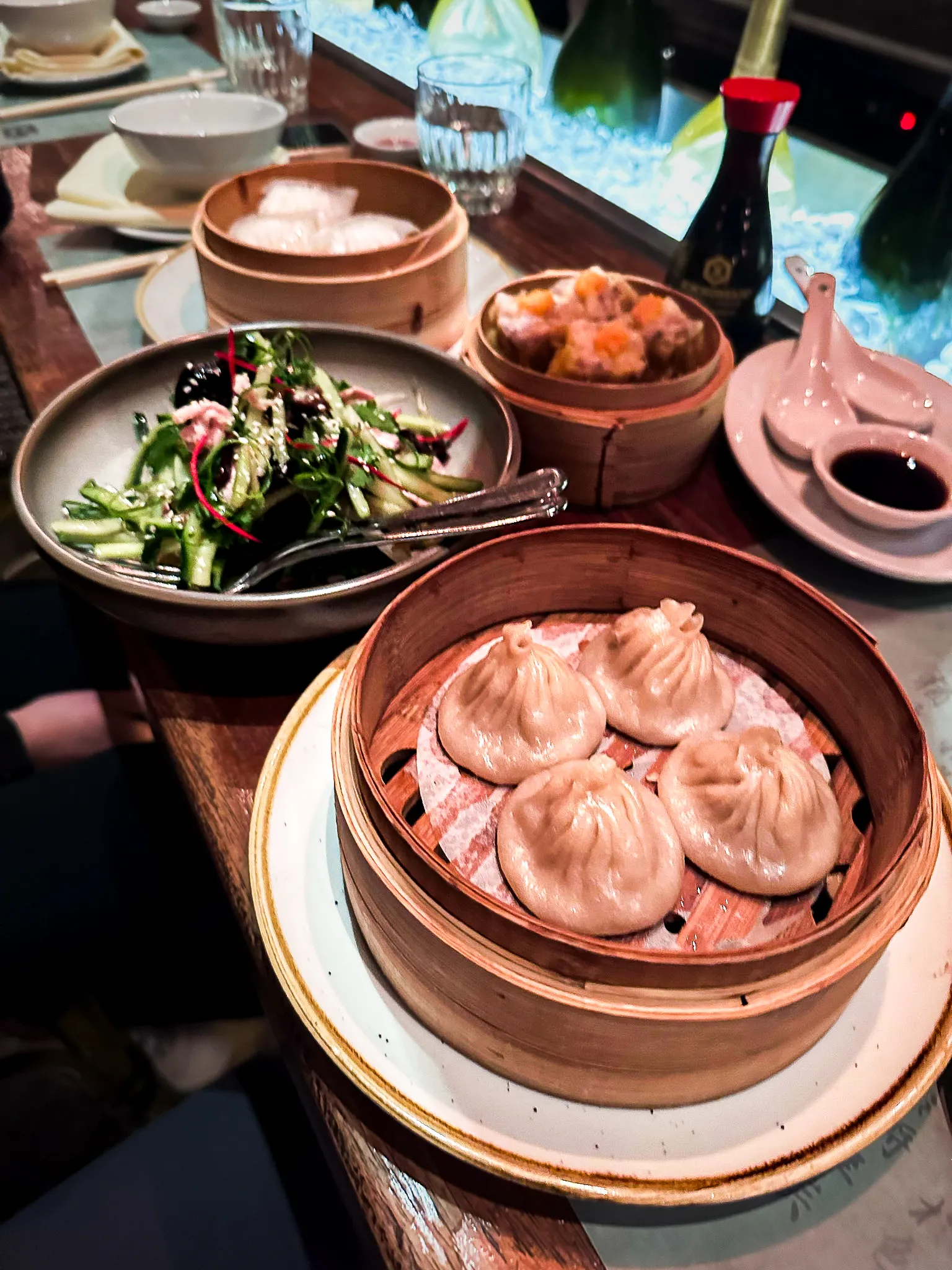 Bamboo steamers of soup dumplings and siu mai on a restaurant table, with a bowl of smashed cucumber salad and a dish of black vinegar
