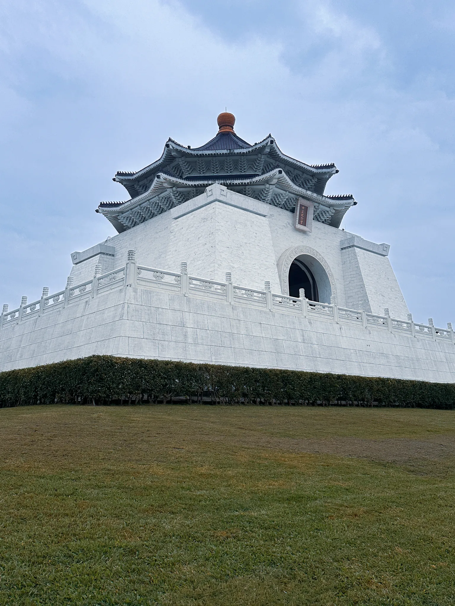 The same hall from the corner of the lawn, looking up past a hedge at the white terrace and the eaves stacked against a pale sky