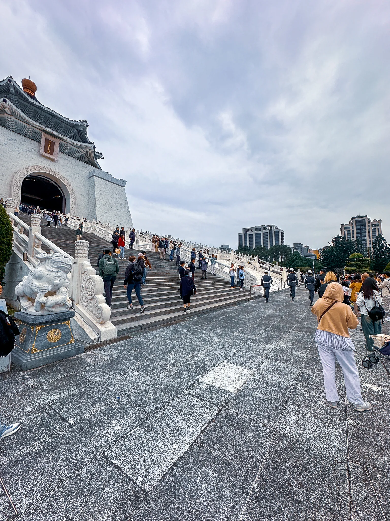 A carved white lion at the foot of the memorial's side stairs, visitors climbing them in winter coats and a woman in a tan hood watching from the paving