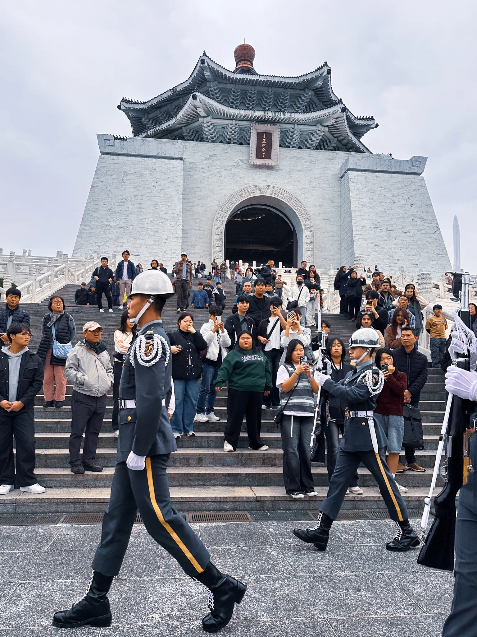 Honour guards in white helmets and gold-striped trousers marching across the foot of the steps, a crowd packed up the stairs behind them holding out phones
