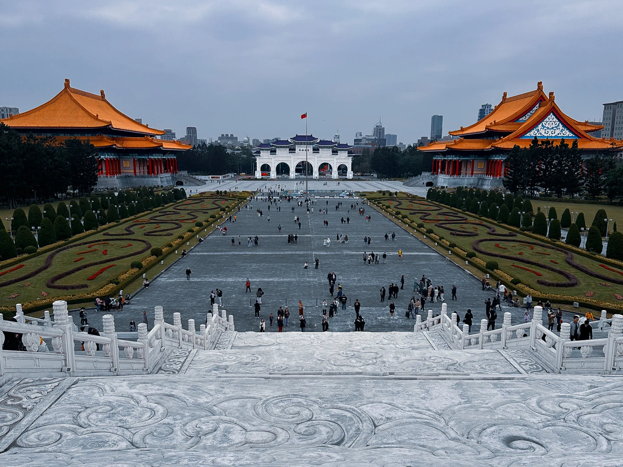 The whole square from the top of the memorial steps, an orange-roofed hall standing to either side and the white gate small at the far end, carved cloud patterns running across the stone underfoot