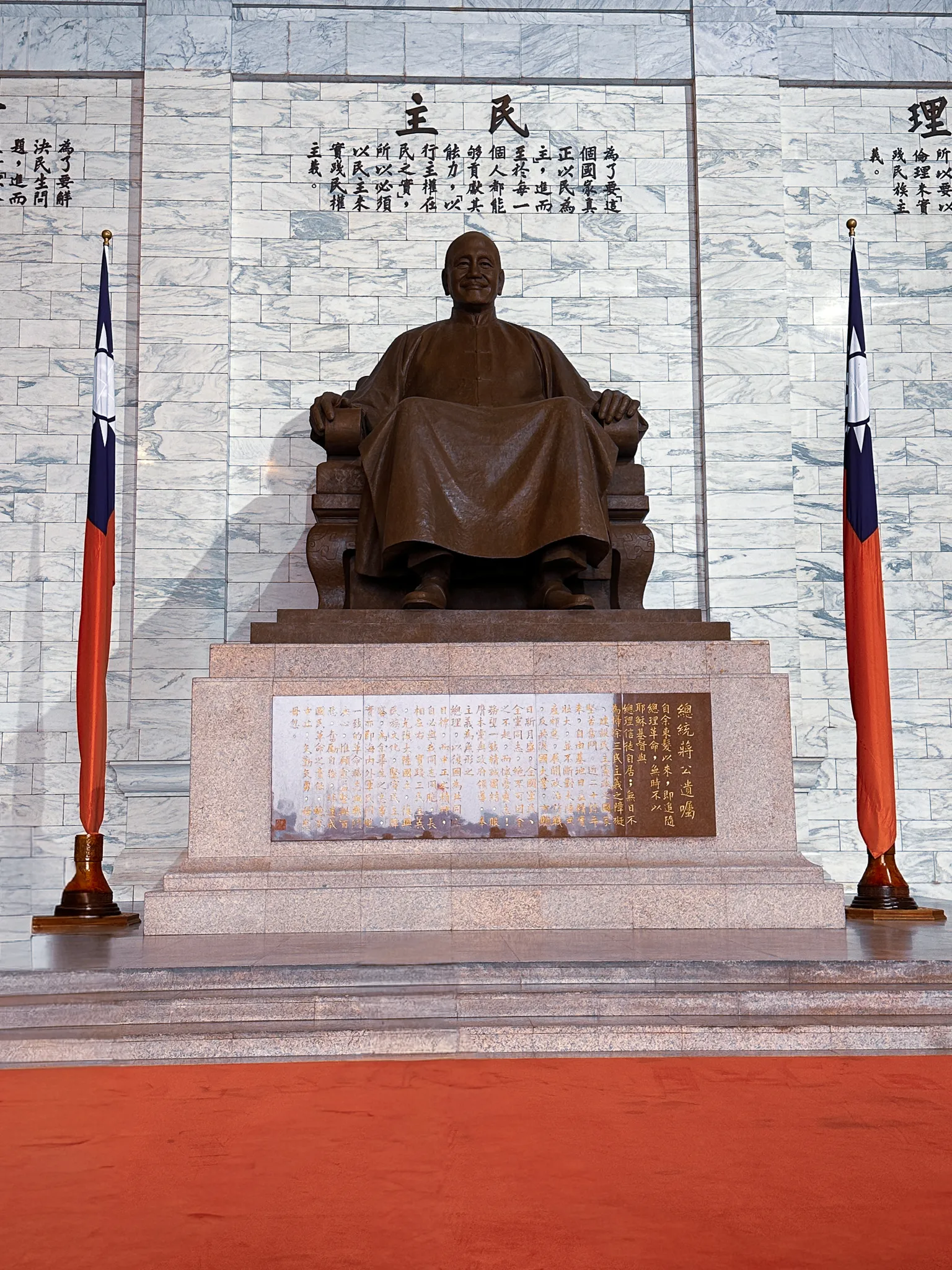 The bronze seated figure inside the hall, a hanging flag to either side and a wall of carved characters behind, a red carpet running up to the plinth