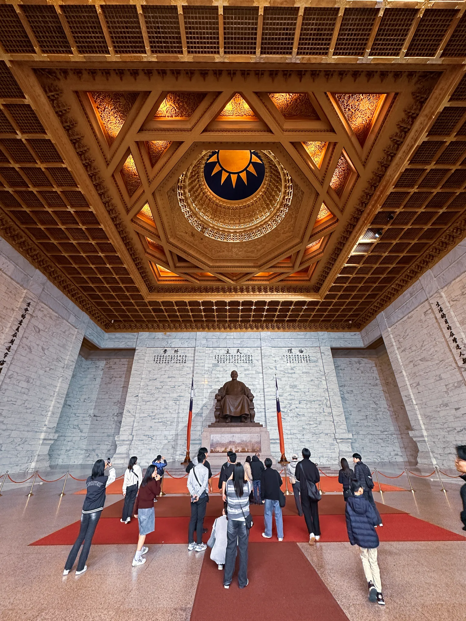 The gilded octagonal ceiling opening above the hall, a blue and white sun at its centre, visitors standing small behind a rope at the statue's feet