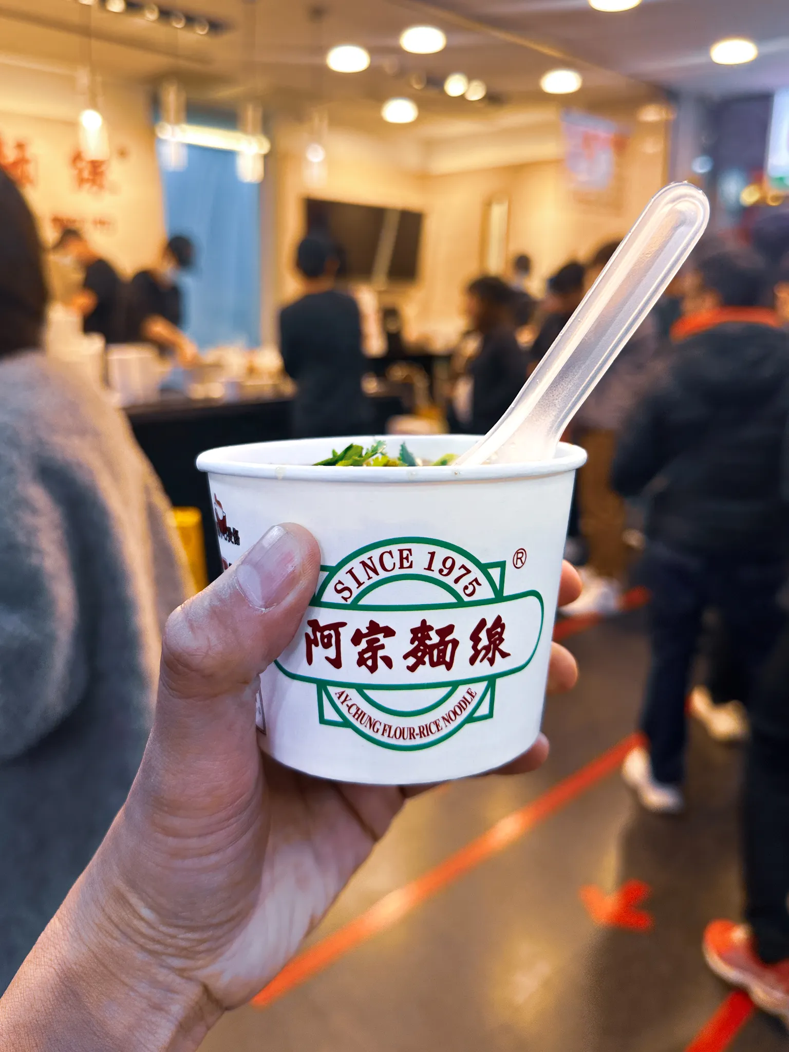 A paper cup of Ay-Chung flour-rice noodles held up in one hand, coriander heaped on top and a plastic spoon standing in it, the counter and the queue blurred behind