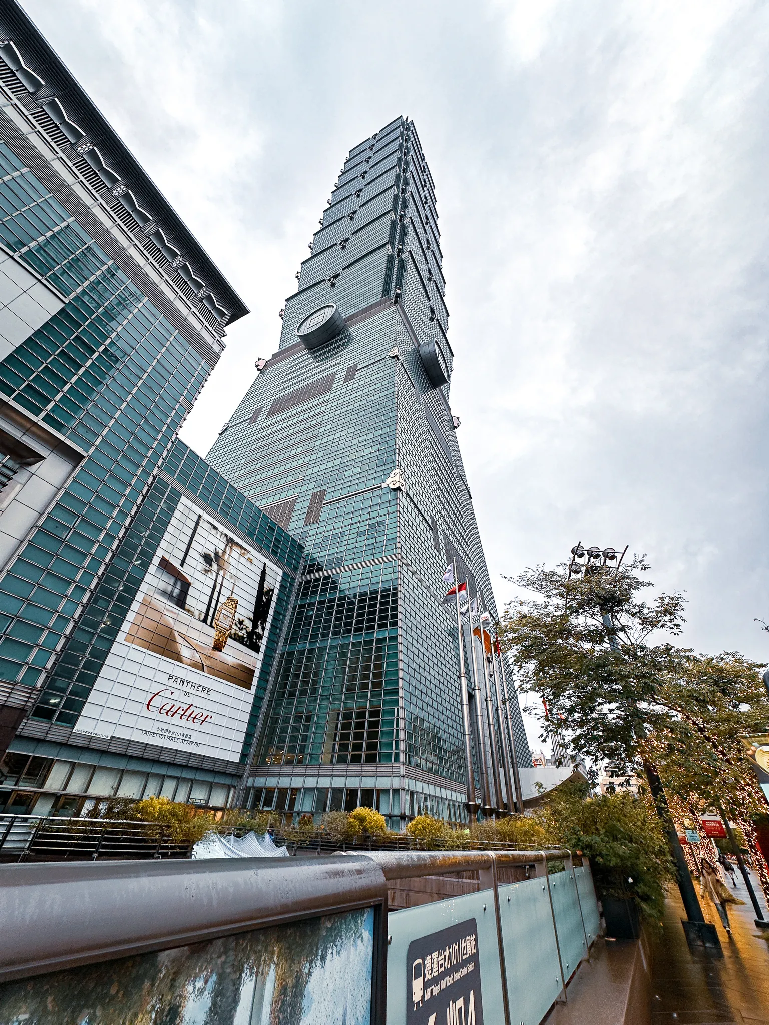Taipei 101 from the pavement, leaning back into flat white cloud with a Cartier billboard on the mall below it and a wet handrail across the foreground