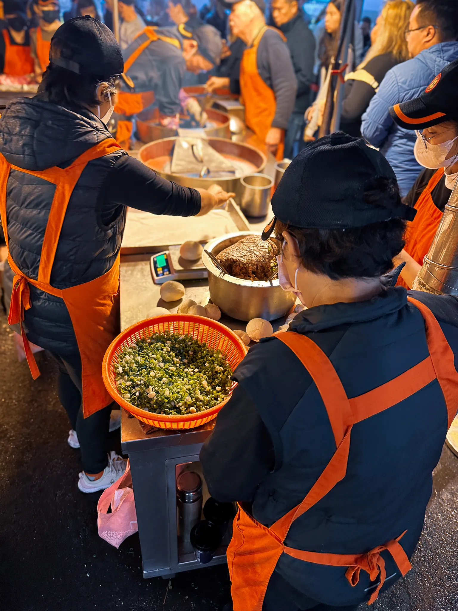 A night market stall mid-service, cooks in orange aprons weighing a basket of chopped spring onion beside a pot of dark filling and a tray of pale dough balls