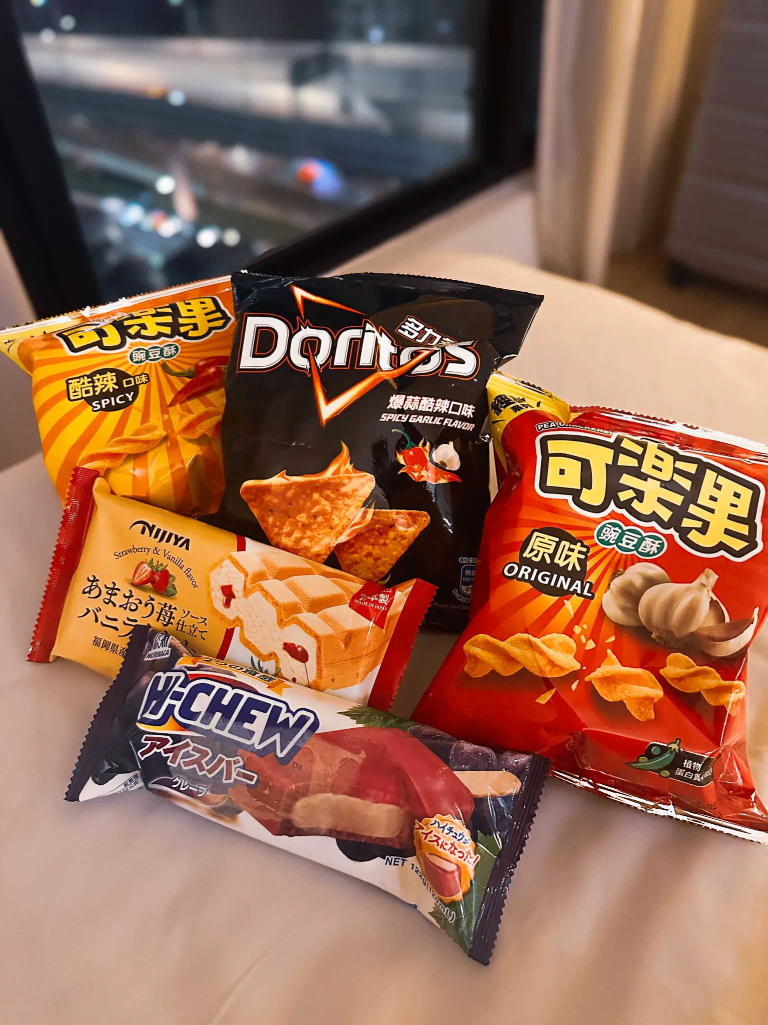 Convenience store snacks spread across a hotel bed - two bags of pea crackers, spicy garlic Doritos, a strawberry sponge and a Hi-Chew ice bar - the city lights out of focus behind