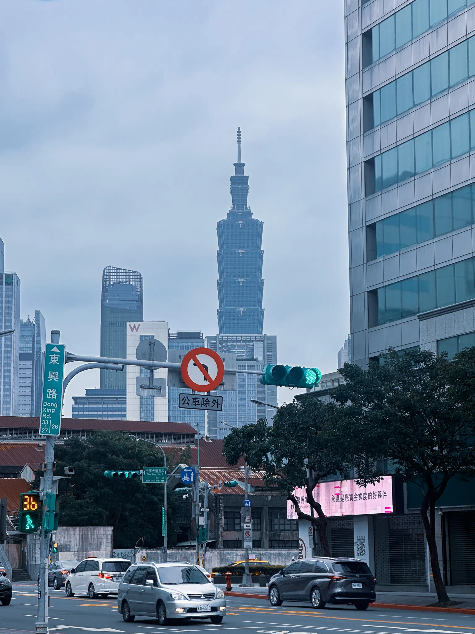 Taipei 101 standing at the end of a street corner, a Dong Xing Rd sign and a no-entry-except-buses roundel in the foreground, traffic waiting under a green light