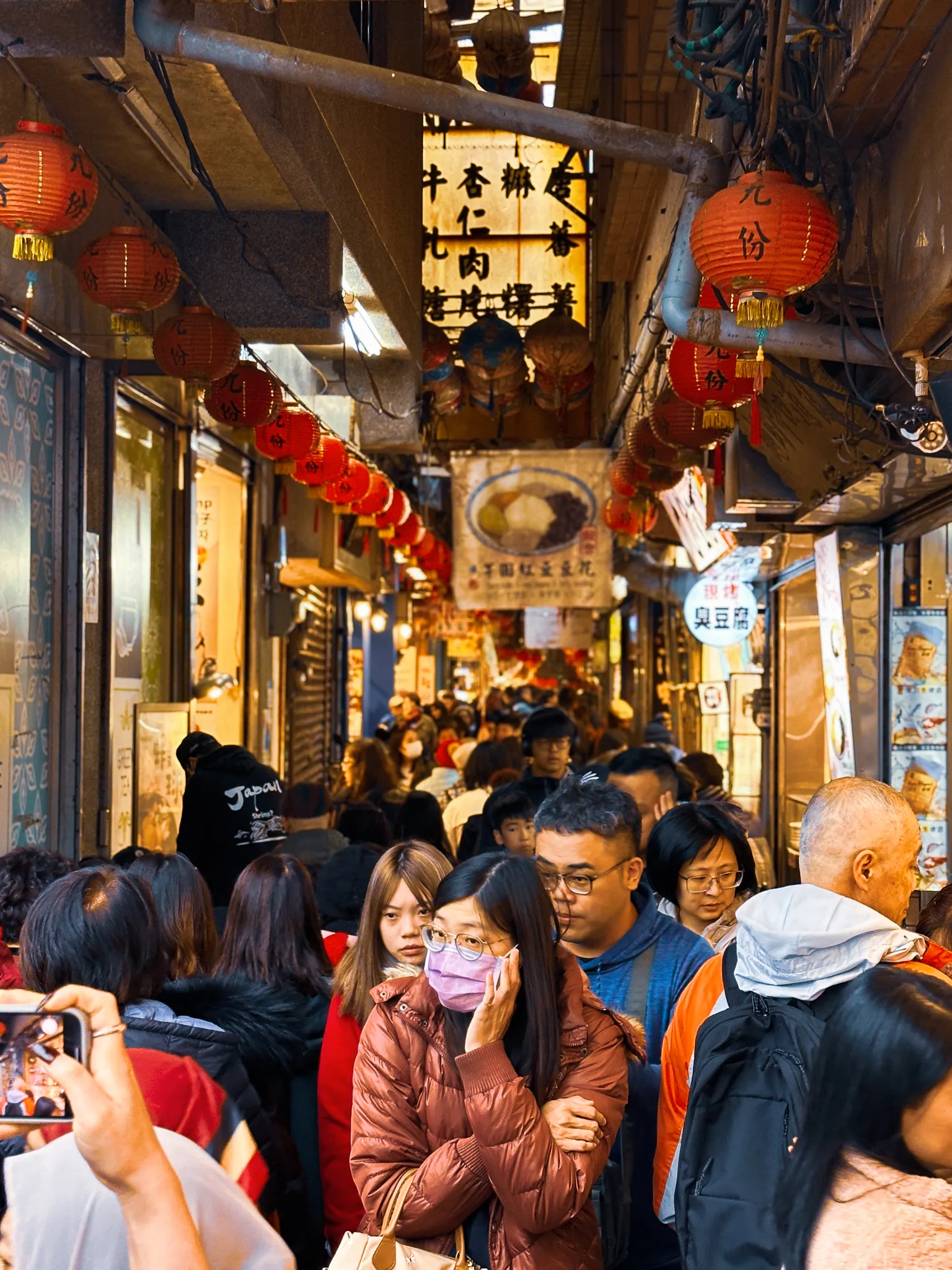 A covered lane packed shoulder to shoulder, rows of red lanterns and hand-painted food signs hung low over the crowd