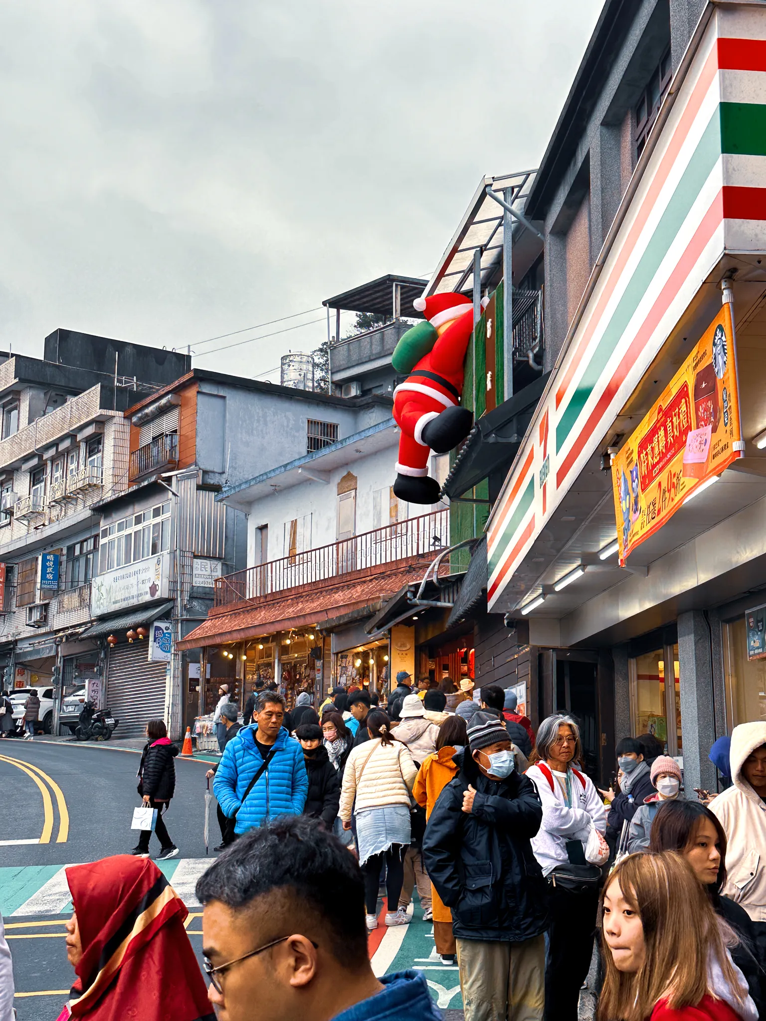 An inflatable Santa climbing the corner of a convenience store on a hill street, the crowd flowing past below and tiled rooftops running down behind