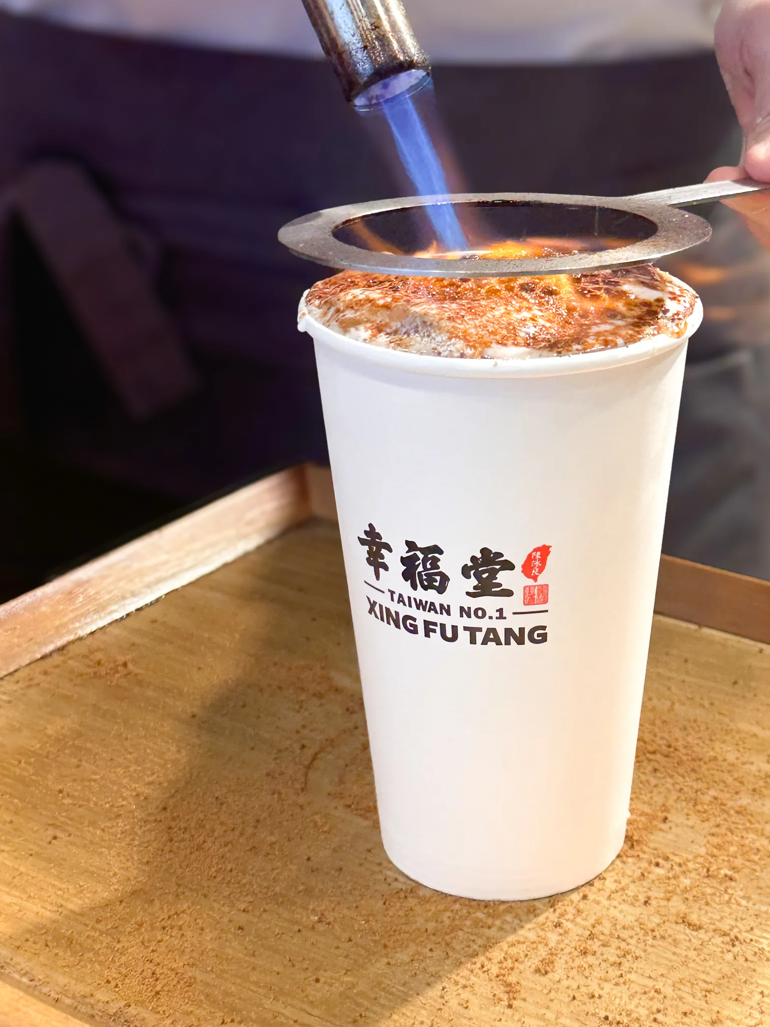 A blowtorch held over the top of a paper cup, the sugar crust catching alight, the cup sitting on a wooden tray dusted brown